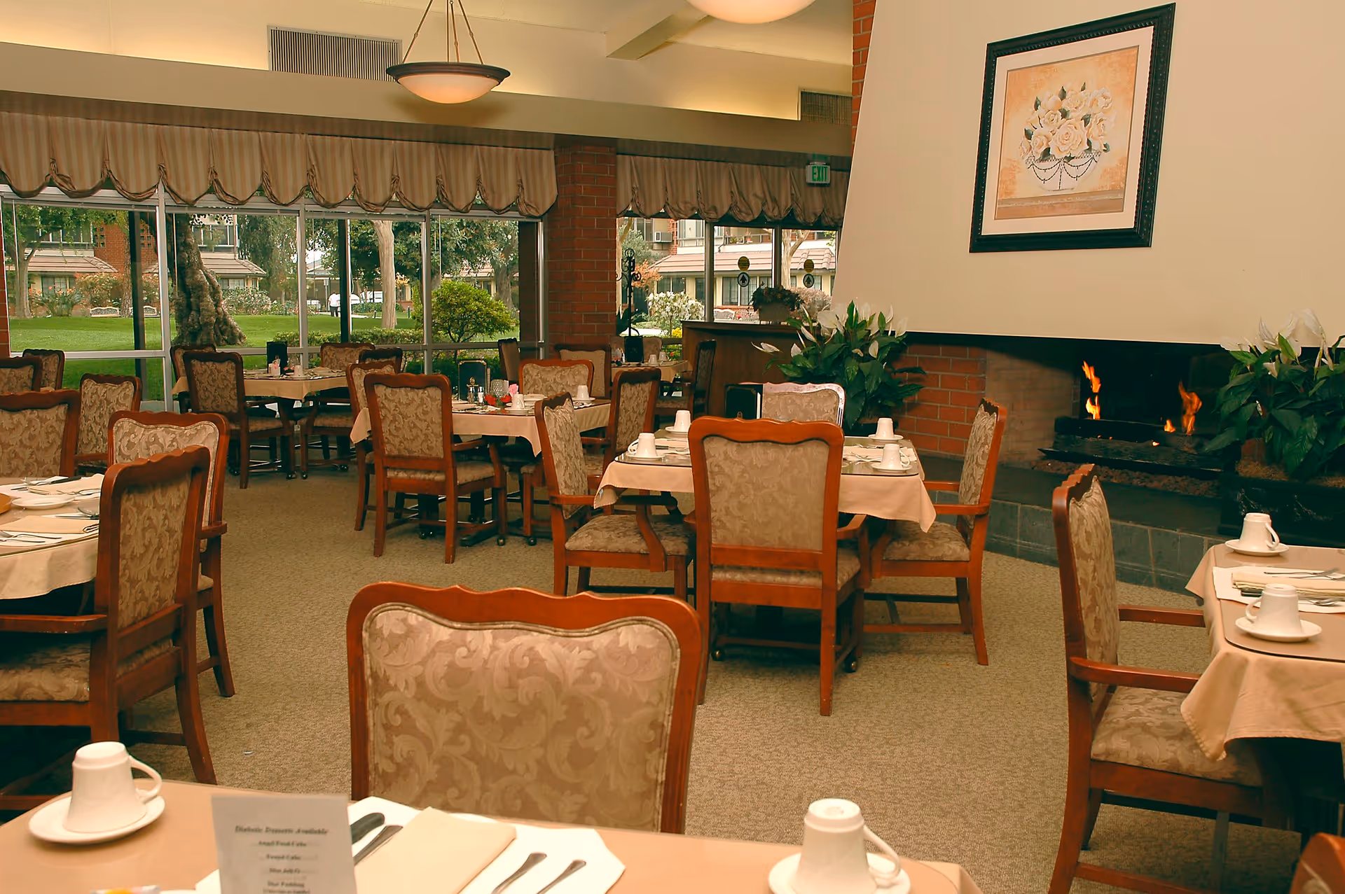 A dining room with multiple tables covered with beige tablecloths, set with white cups, plates, and silverware. The room features upholstered wooden chairs, a fireplace with a fire burning, framed floral artwork above the fireplace, large windows with a view of a garden outside, and plants near the fireplace.