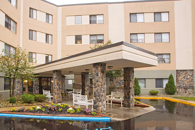 Exterior view of a multi-story senior living facility with a covered entrance supported by stone pillars. White benches are placed under the entrance canopy, and there are landscaped flower beds and small trees around the driveway.