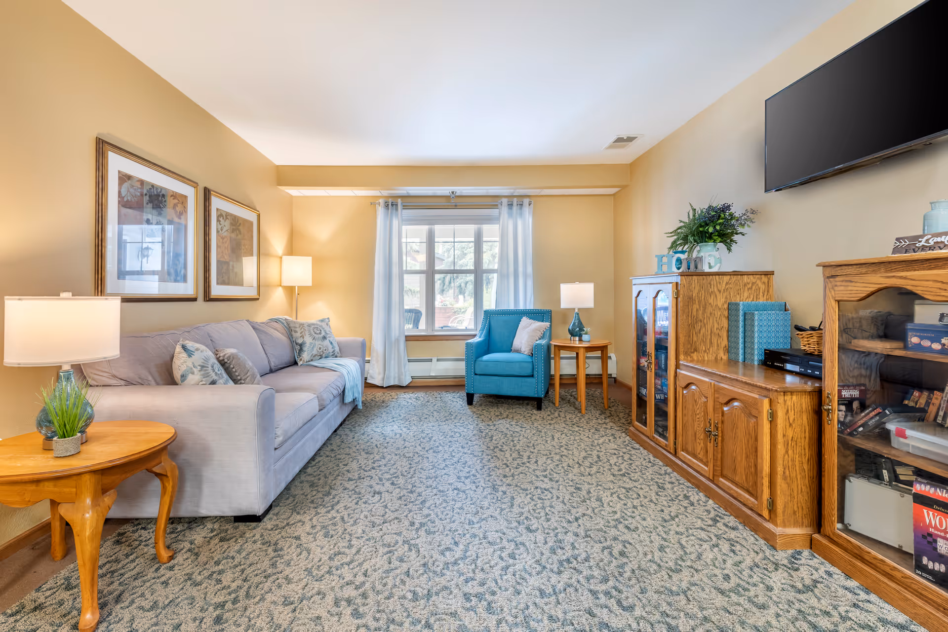 A cozy living room with a light gray sofa adorned with patterned pillows on the left, a wooden side table with a lamp and a small plant, a blue armchair with a pillow near a window with white curtains, and wooden cabinets on the right holding various items. A flat-screen TV is mounted on the wall above the cabinets. The room has beige walls and a patterned carpet.