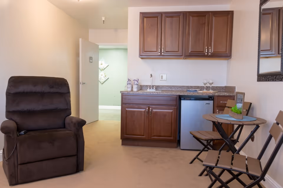 Interior view of a room in Casa De Flores featuring a brown recliner chair on the left, a kitchenette with wooden cabinets, a small sink, and a mini fridge in the center, and a small round table with two folding chairs on the right. A mirror hangs on the wall above the table, and the door to another room is open, showing a green wall with decorative white flowers.