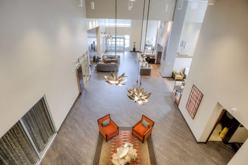 A spacious and modern senior living facility common area with high ceilings, two contemporary pendant lights, two orange armchairs around a decorative table on a patterned rug, and additional seating areas with sofas and chairs in the background near large windows.