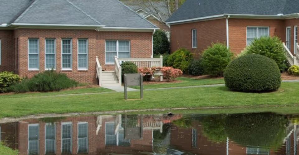 Exterior view of a brick building with multiple windows and small staircases leading to entrances, surrounded by well-maintained bushes and greenery, reflected in a calm pond in the foreground.