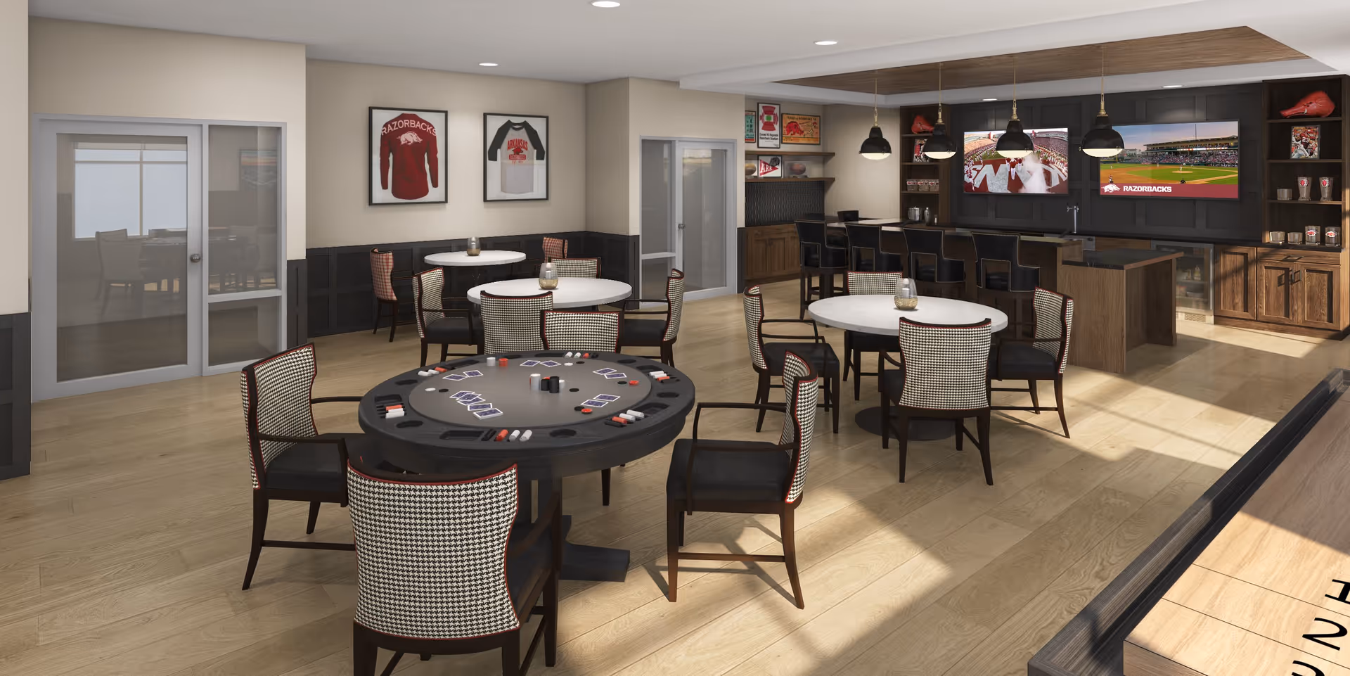 A spacious recreational room with multiple round tables and chairs, including a poker table with playing cards and chips. The room features a bar area with high stools, two large flat-screen TVs mounted on the wall displaying sports content, and shelves with decorative items. The walls have framed sports jerseys, and the floor is light wood. There is a glass door leading to another room with a conference table and chairs.