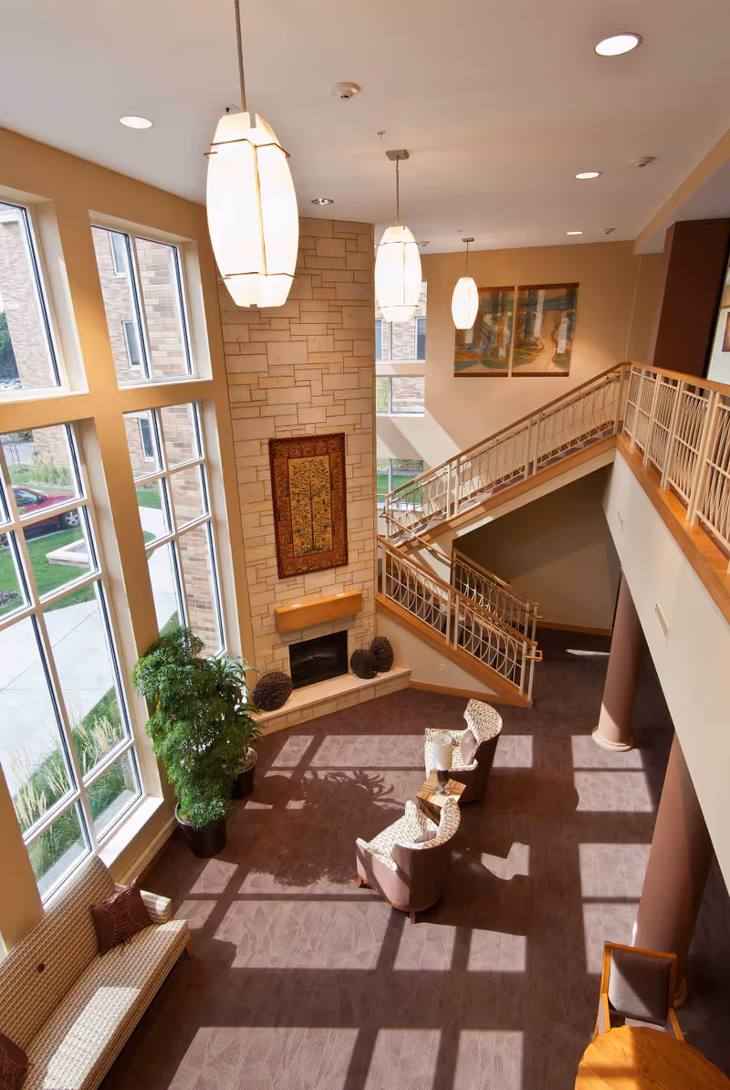 A bright and spacious living area with large floor-to-ceiling windows letting in natural light. The room features a stone fireplace with decorative artwork above it, two hanging pendant lights, a staircase with a wooden handrail, and several chairs arranged around a small table. There is also a potted plant and a cushioned bench near the windows.