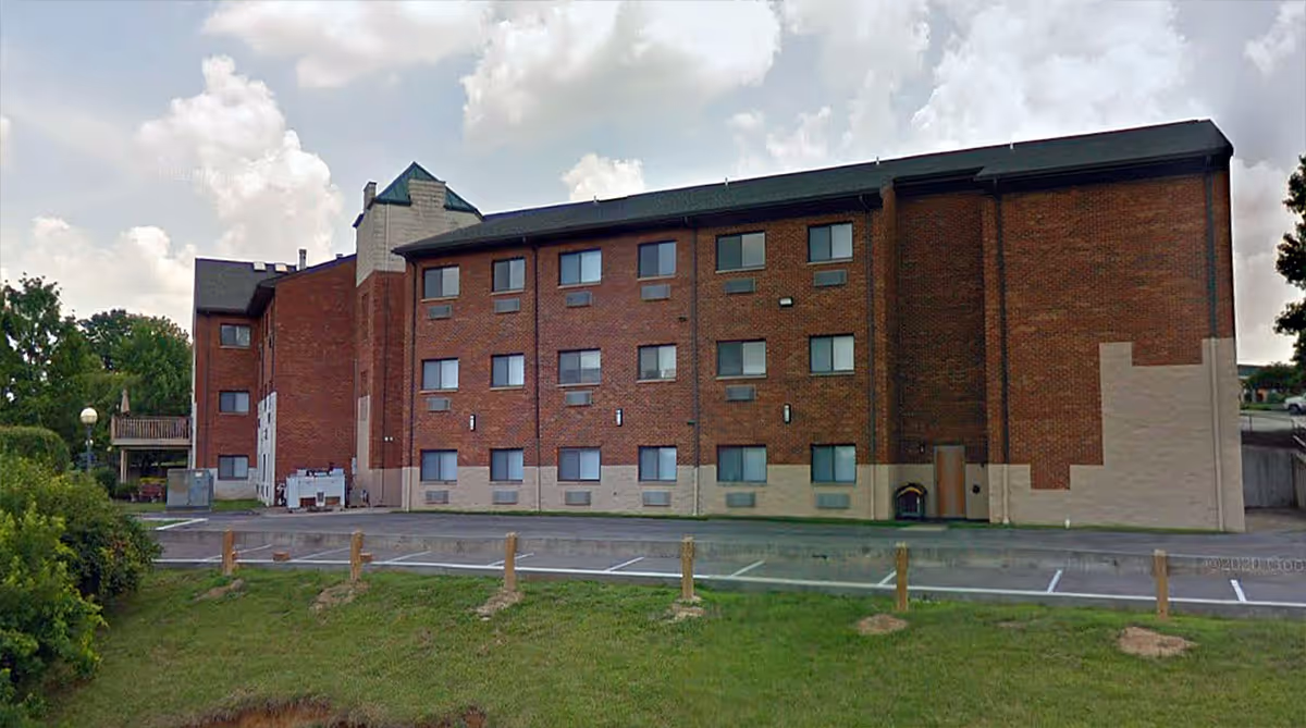 Exterior view of a three-story brick building with multiple windows, a small parking area in front, and a grassy area with wooden posts. The sky is partly cloudy.