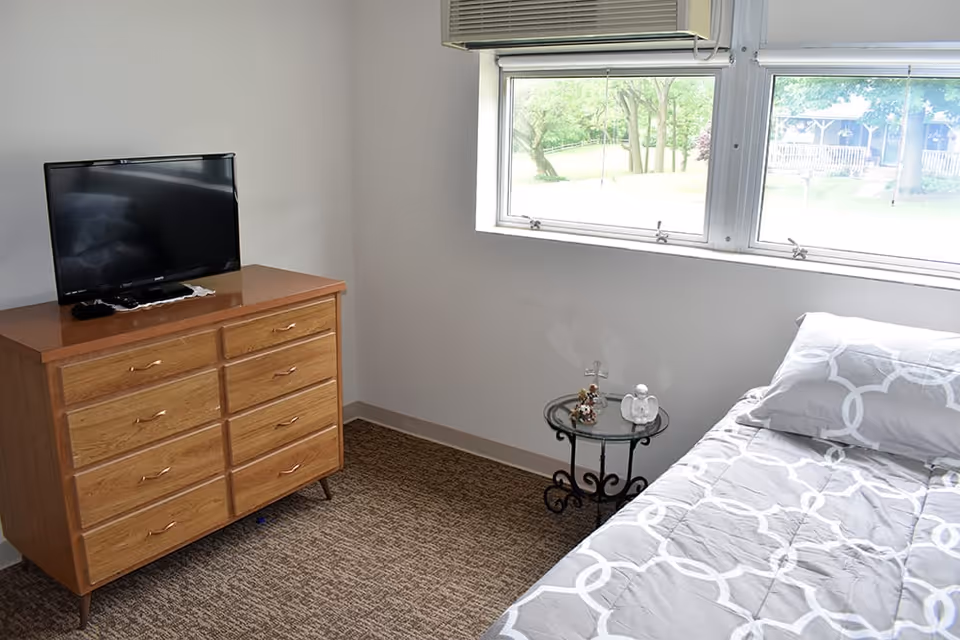 A small bedroom with a bed covered in a gray and white patterned comforter and pillow. Next to the bed is a round glass-top side table with decorative items on it. Across from the bed is a wooden dresser with six drawers, on top of which sits a flat-screen TV. A large window with a view of green trees and grass lets in natural light. An air conditioning unit is installed above the window.