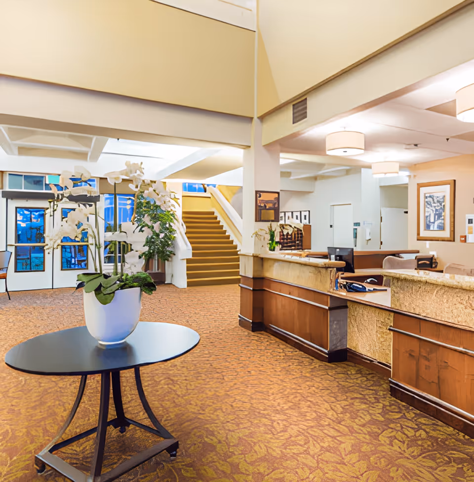 Interior view of a senior living facility lobby with a round table holding a white orchid plant in the foreground, a reception desk to the right, carpeted floor with a floral pattern, and a staircase leading to an upper floor in the background.