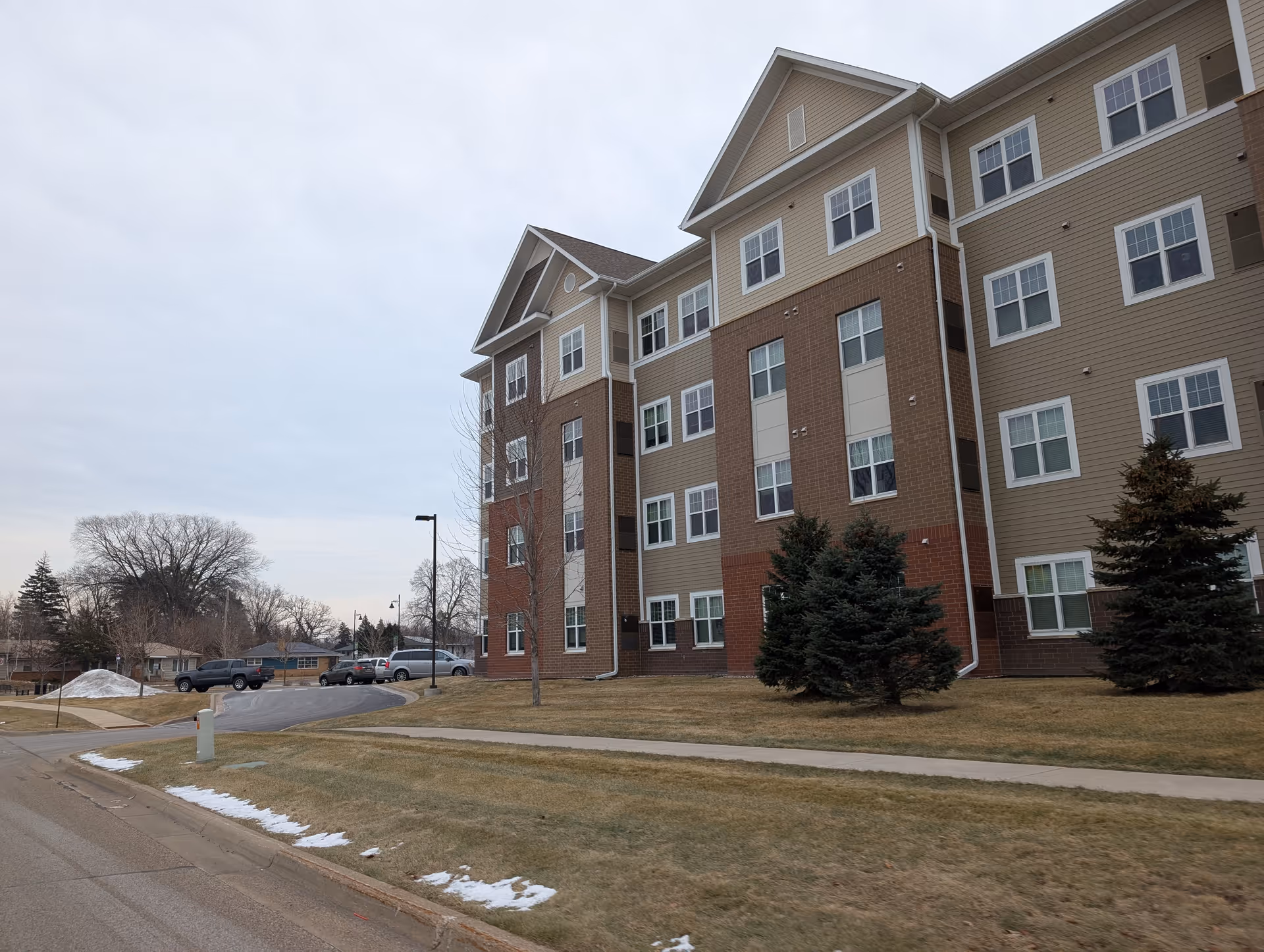 Exterior view of a multi-story senior living facility building with beige and brick facade, several windows, small evergreen trees, and a parking area with cars. The sky is overcast and patches of snow are visible on the grass and sidewalk.