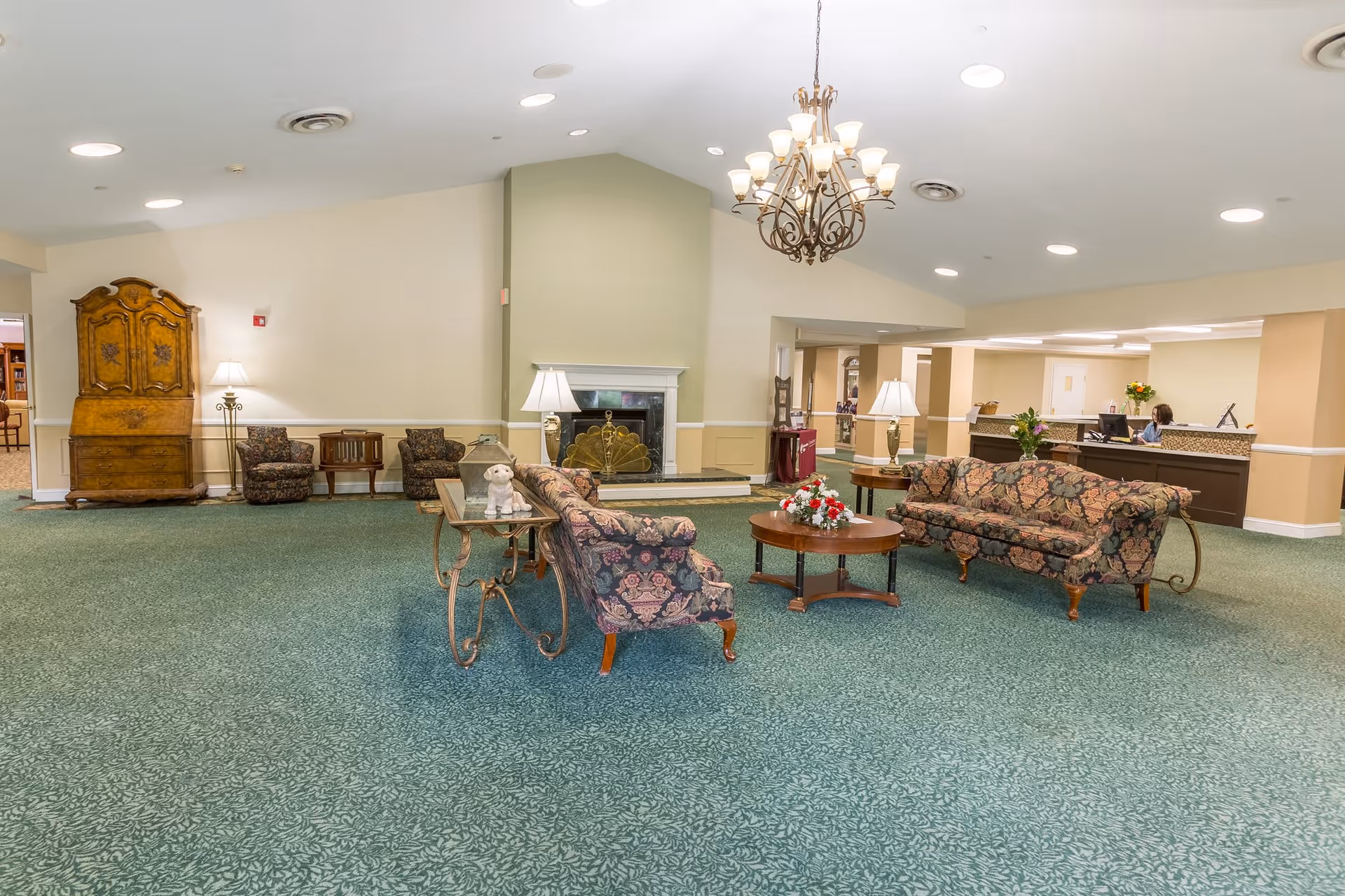 A spacious senior living facility common area with green patterned carpet, floral upholstered sofas and armchairs arranged around a wooden coffee table with a flower arrangement. There is a fireplace with a decorative screen, a chandelier hanging from the ceiling, and a reception desk with a staff member seated behind it. The room is softly lit with table lamps and ceiling lights, and there is an antique wooden cabinet and additional seating along the walls.
