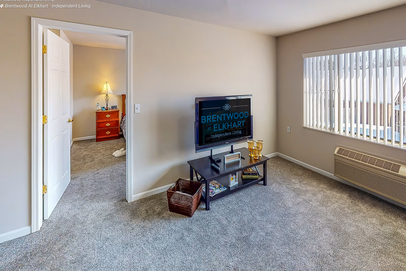 A bright living room with beige walls and carpeted floor featuring a flat-screen TV on a black stand with decorative items and a basket underneath. A large window with vertical blinds lets in natural light. An open door leads to a bedroom with a wooden nightstand, lamp, and slippers on the carpet.
