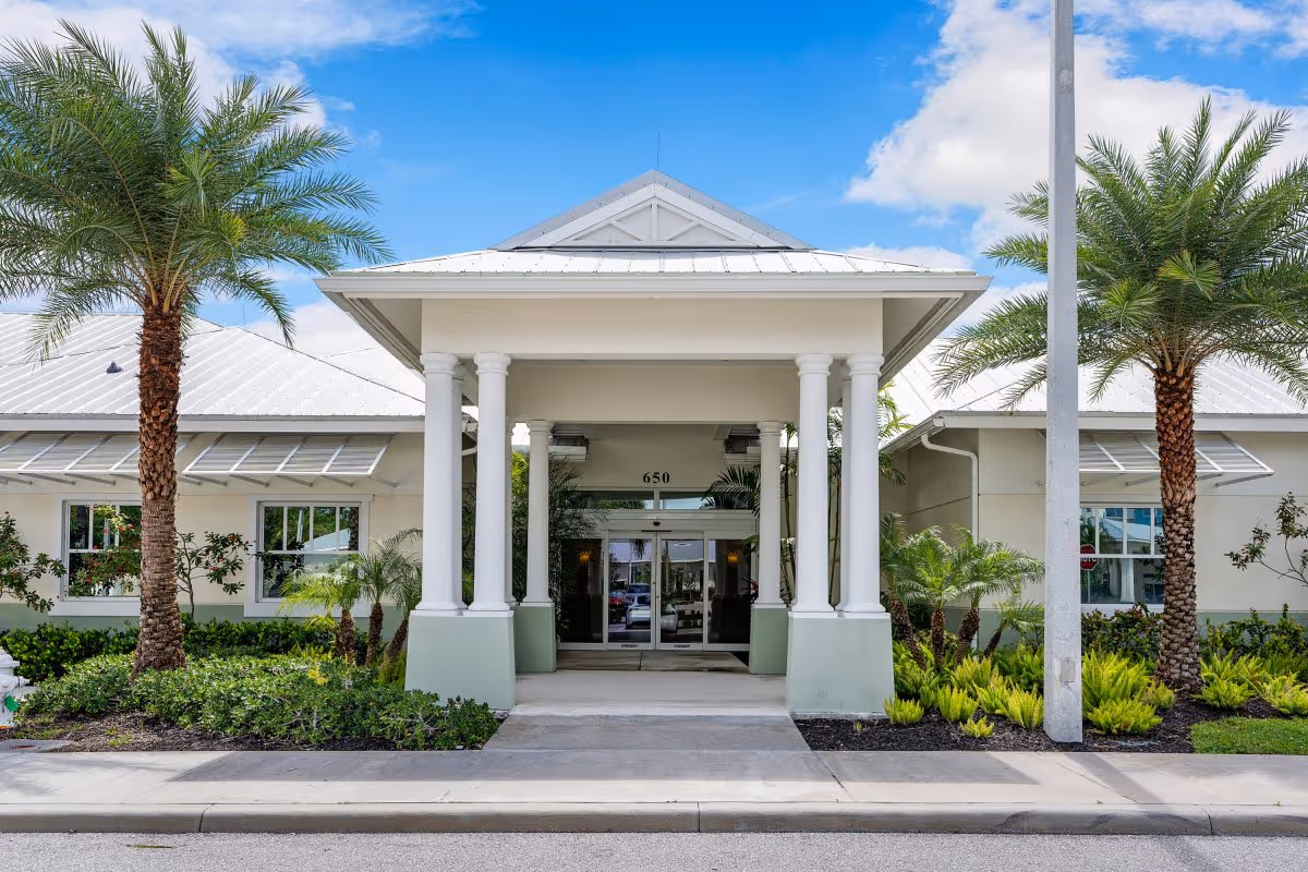 Front entrance of a senior living facility with white columns, a covered walkway, palm trees, and well-maintained landscaping under a blue sky with some clouds.