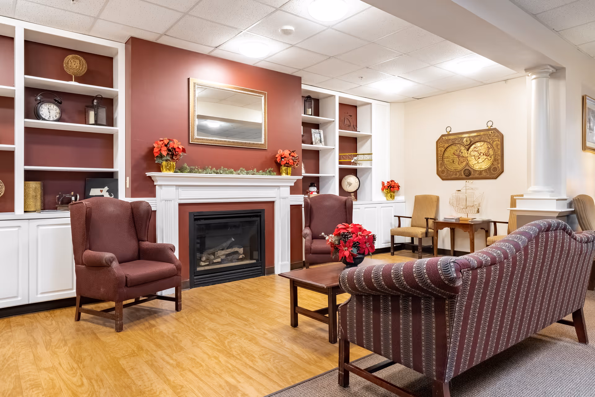 A cozy senior living facility common area with a fireplace centered on a red accent wall, flanked by white built-in shelves and cabinets. The room features two maroon armchairs, a striped sofa, a wooden coffee table with a red floral arrangement, and additional chairs and tables. Decorative items include a mirror above the fireplace, a vintage clock, globe, and wall art depicting an antique map.