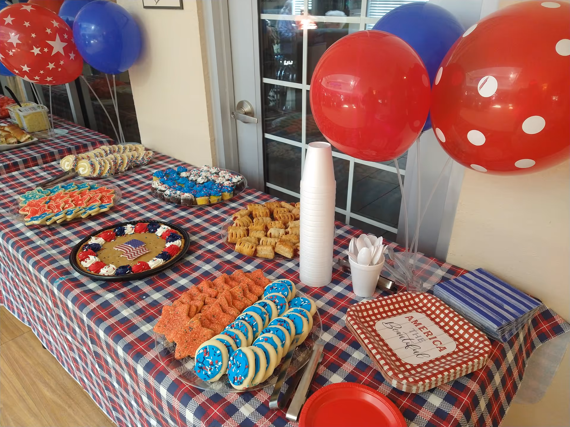 A table covered with a red, white, and blue plaid tablecloth displaying various patriotic-themed cookies, cupcakes, and pastries. There are red, blue, and white balloons tied to the table, along with stacks of disposable cups, plastic utensils, napkins, and plates with the phrase 'America the Beautiful'. The setting appears to be indoors near a glass door.