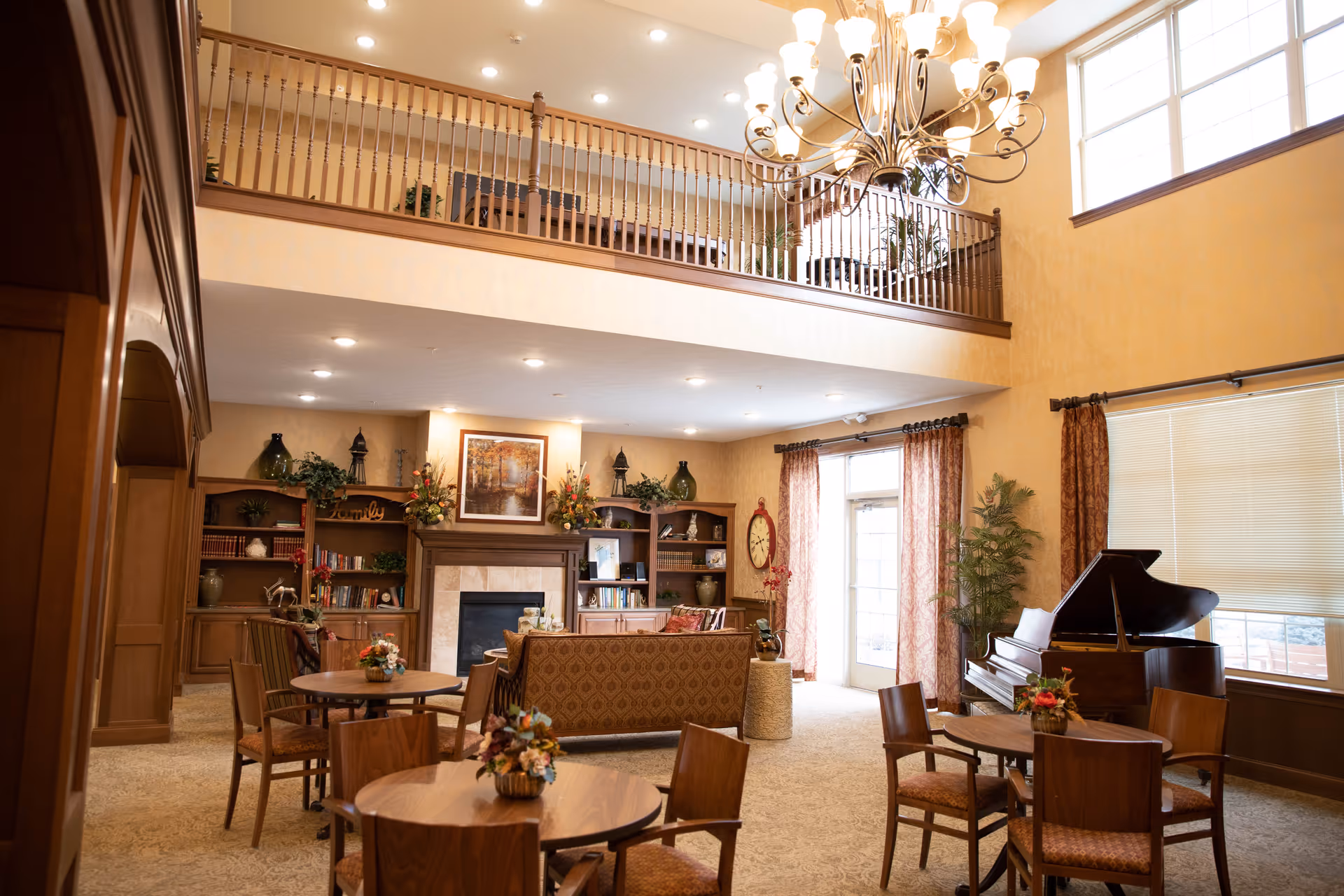 A spacious and warmly decorated living room area in Forest Hills Commons featuring a high ceiling with a chandelier, a balcony railing above, multiple round wooden tables with chairs, a grand piano near large windows with curtains, a fireplace surrounded by built-in wooden shelves filled with books and decorative items, and floral arrangements on the tables and shelves.