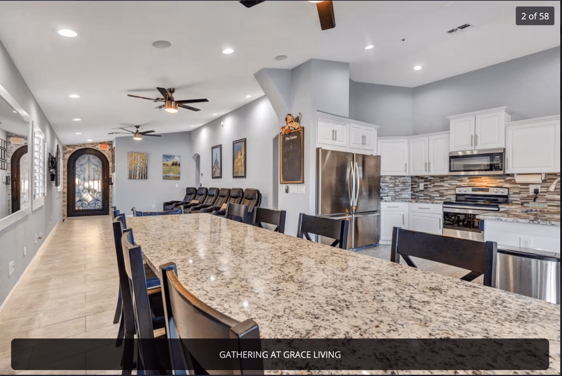 Interior view of a senior living facility showing a long granite countertop table with several black chairs around it. In the background, there is a modern kitchen with white cabinets, stainless steel appliances including a refrigerator, stove, and microwave. The room has light gray walls, recessed ceiling lights, ceiling fans, and several framed pictures on the walls. There is a decorative arched door with wrought iron detailing at the far end of the room.
