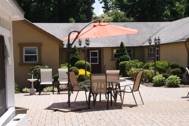 Outdoor patio area with a round table and six chairs under a large orange umbrella. The patio is paved with bricks and surrounded by bushes and small trees. The building in the background has beige walls and multiple windows.
