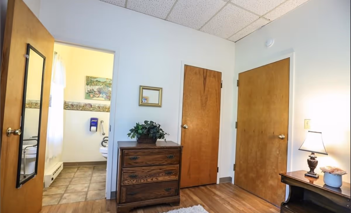 Small furnished bedroom with a wooden dresser and nightstand, two closed doors, and an open doorway showing a bathroom.