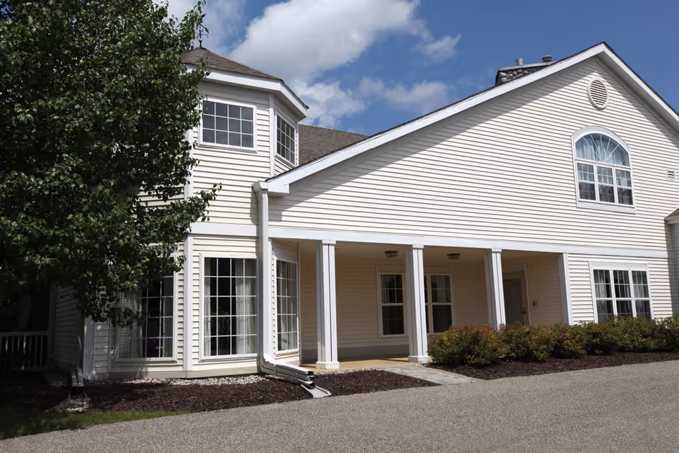 Exterior view of a light-colored building with multiple windows, a covered porch supported by white columns, and a tree on the left side under a partly cloudy sky.