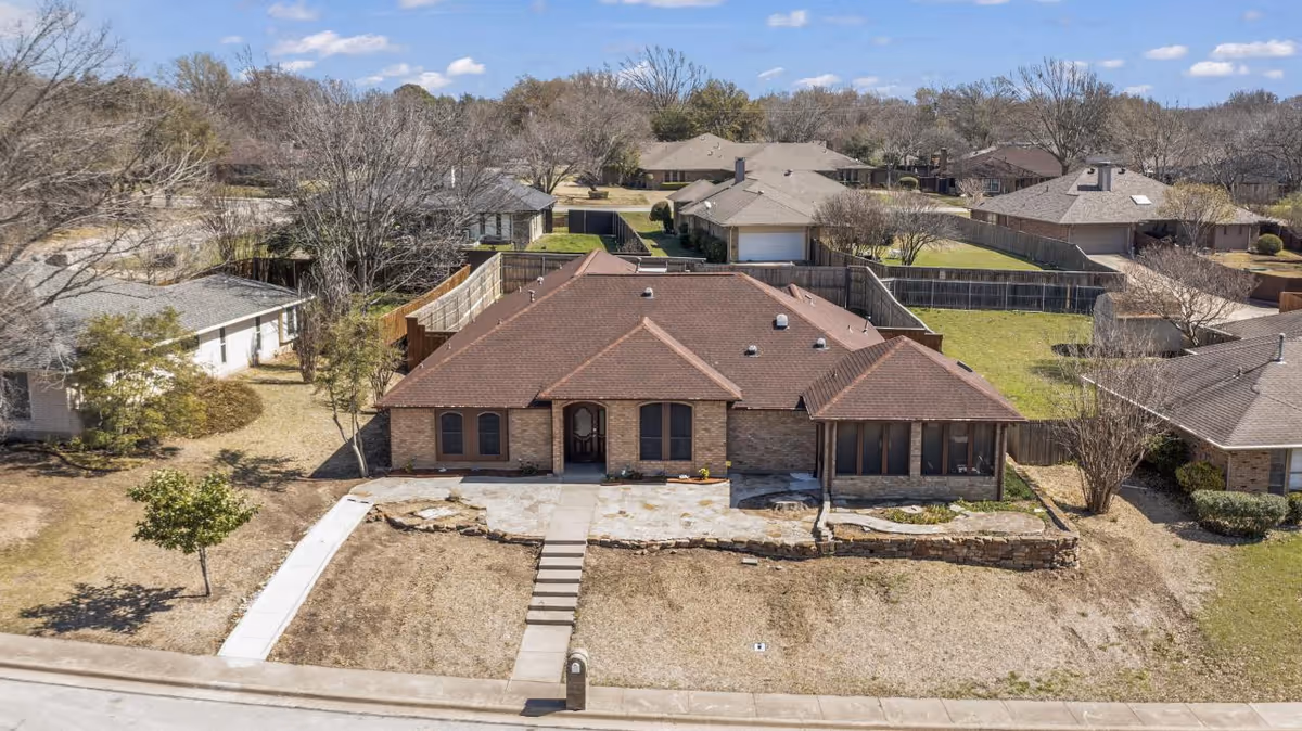 Aerial view of a single-story brick house with a brown roof, a concrete walkway leading to the front door, and a dry lawn with minimal landscaping. The house is situated in a suburban neighborhood with similar houses and leafless trees under a partly cloudy blue sky.