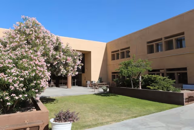 Outdoor courtyard with grass, potted plants, flowering trees and patio furniture in front of a two-story tan building.