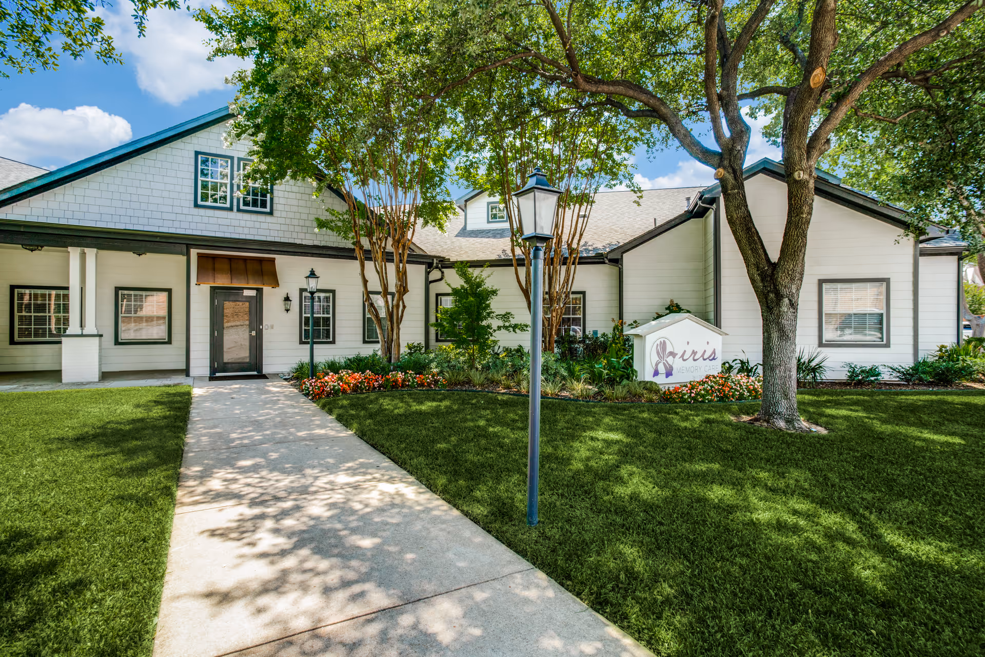 Exterior view of a single-story white building with green trim, surrounded by well-maintained grass, trees, and flower beds. A concrete walkway leads to the entrance door, and there is a lamp post along the path. A sign near the building reads 'iris'.