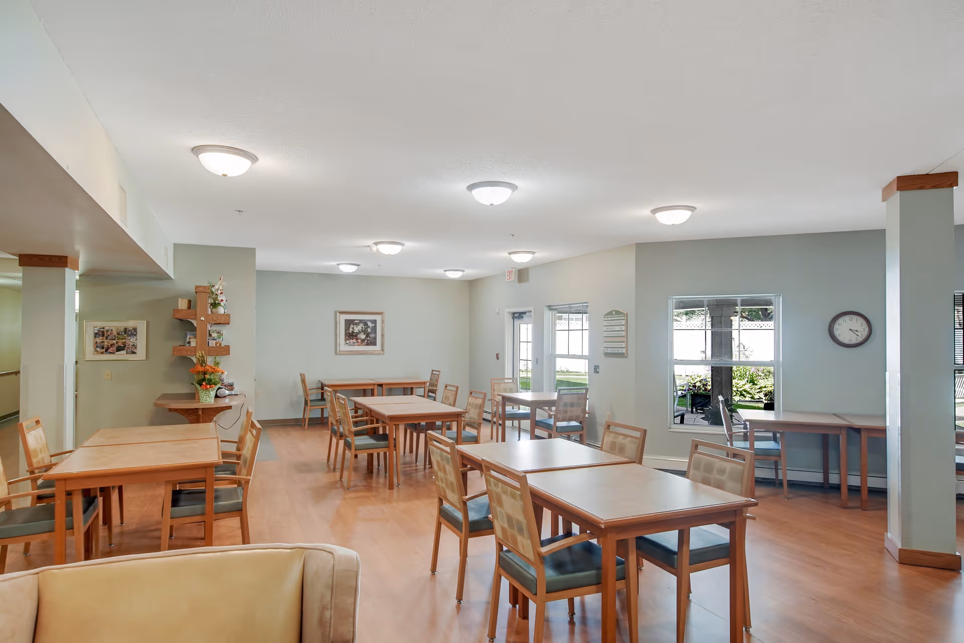A bright and clean dining room in Oak Ridge Assisted Living with multiple wooden tables and chairs arranged neatly. The room has light green walls, several ceiling lights, a clock on the wall, and windows letting in natural light. There is a small shelf with flowers and decorations on the left side and a door leading outside at the back.