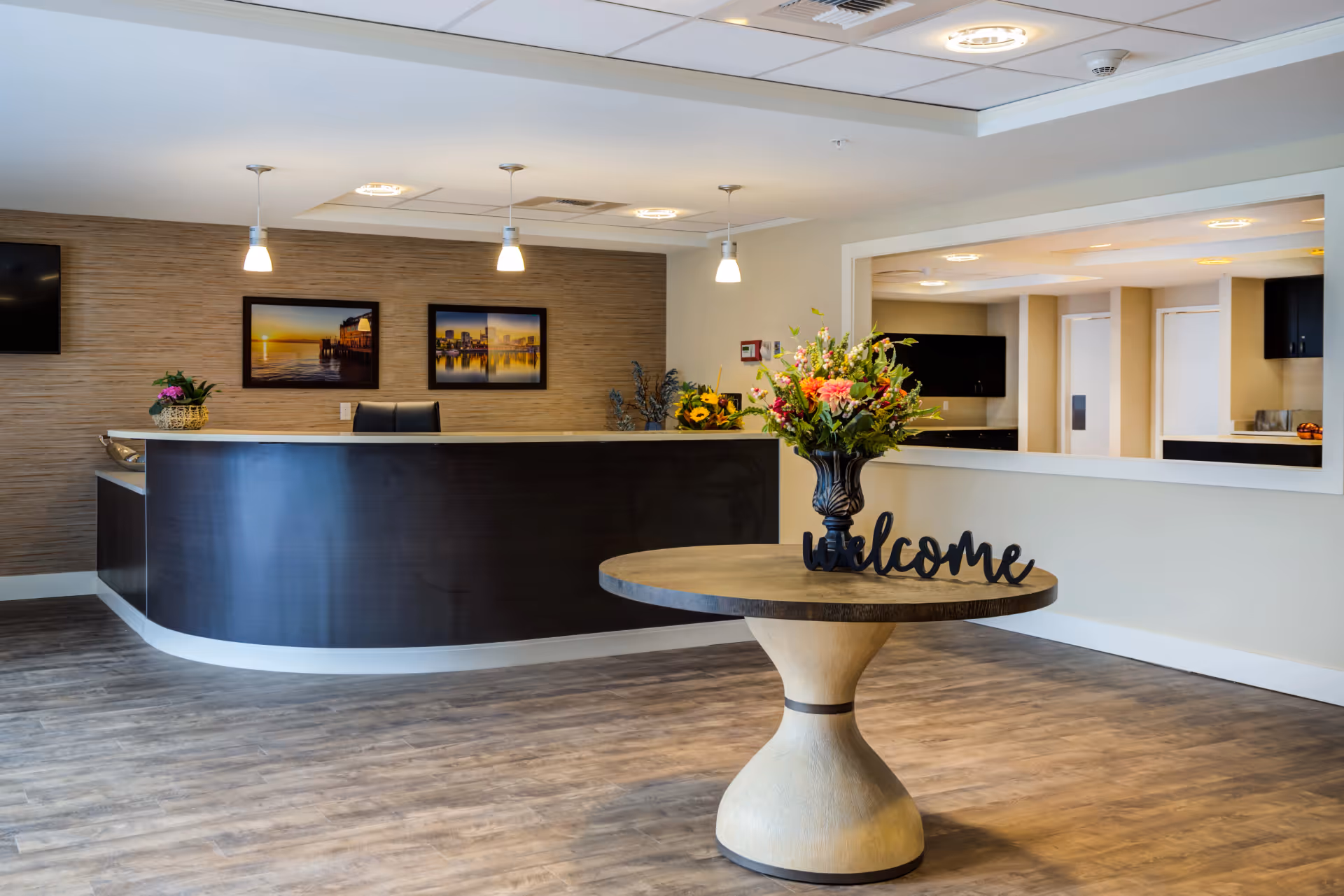 A senior living reception area featuring a curved dark wood front desk, a round table with a vase of flowers and a 'welcome' sign, and framed artwork on the wall.