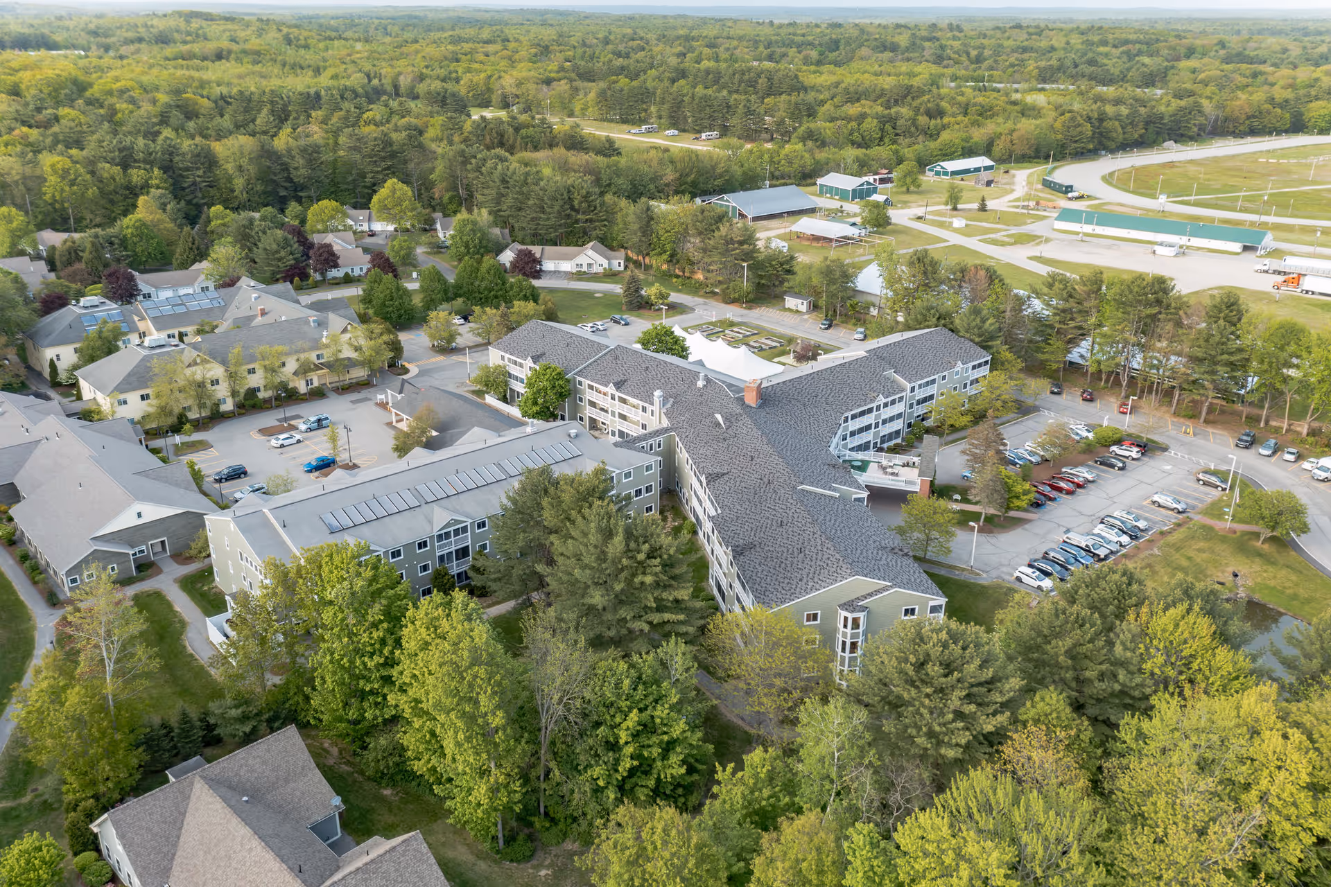 Aerial view of The Highlands senior living facility surrounded by dense green trees and open land. The facility consists of multiple connected buildings with gray roofs and parking lots filled with cars. There are additional smaller buildings and open spaces visible in the background.
