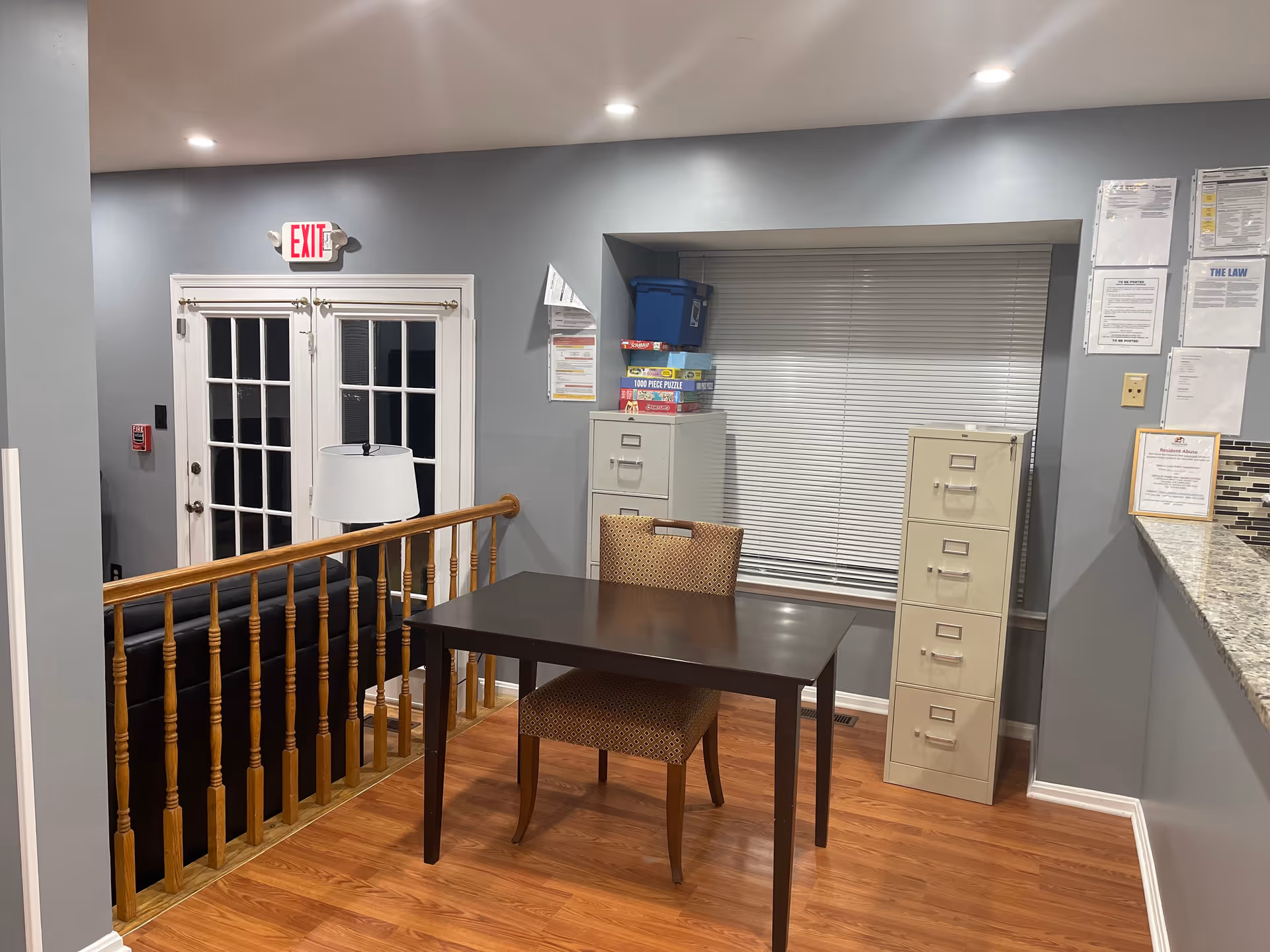 Interior view of a room with gray walls and wooden flooring featuring a dark wooden table with a patterned chair. Behind the table are two beige filing cabinets, a window with closed blinds, and some board games stacked on top of one filing cabinet. To the left, there is a wooden railing overlooking a lower level with a black couch and a white lamp. Double glass doors with an illuminated exit sign above them are visible in the background. Various notices and papers are pinned on the wall near the filing cabinets and counter.