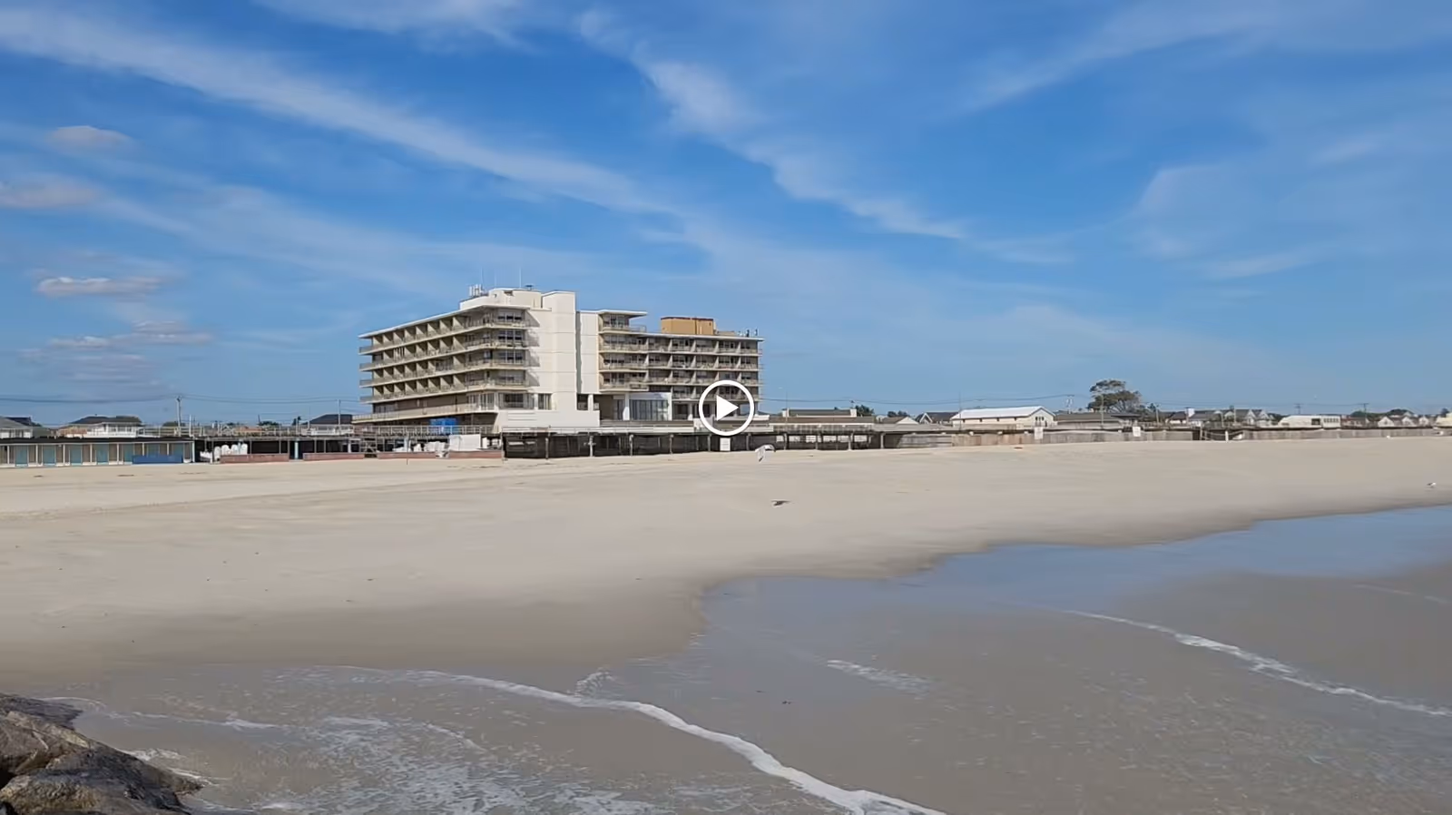 View of a multi-story building near a wide sandy beach with gentle waves under a blue sky with some clouds.