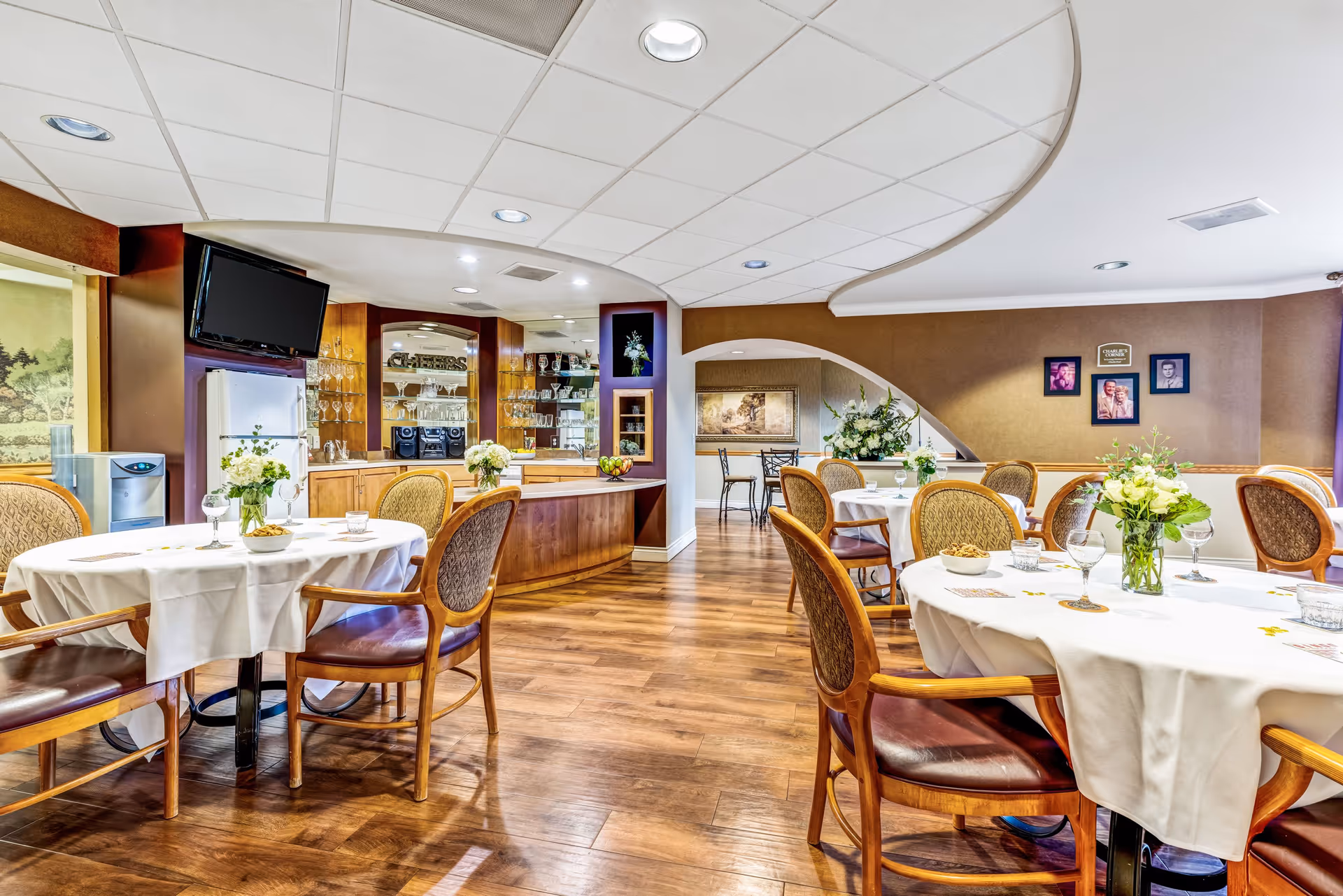 A bright and inviting dining room with round tables covered in white tablecloths, each adorned with floral centerpieces and glassware. Wooden chairs with cushioned seats surround the tables. The room features wood flooring, a curved ceiling design, and a small bar area with glass shelves displaying glassware. A wall-mounted TV and a water cooler are visible in the background, along with framed pictures on the wall.