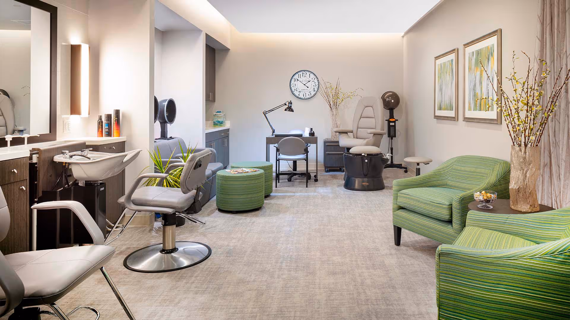 A bright and modern salon area in a senior living facility featuring salon chairs, a hair washing station, a manicure table with a lamp and chair, green striped armchairs, decorative plants, and framed artwork on the walls.