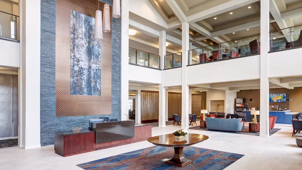 Spacious and modern senior living facility lobby with high ceilings, a large decorative wall art piece behind a wooden reception desk, a round wooden table with a floral centerpiece on a patterned rug, and seating areas with sofas and chairs. The space features an open mezzanine level with glass railings and warm lighting.