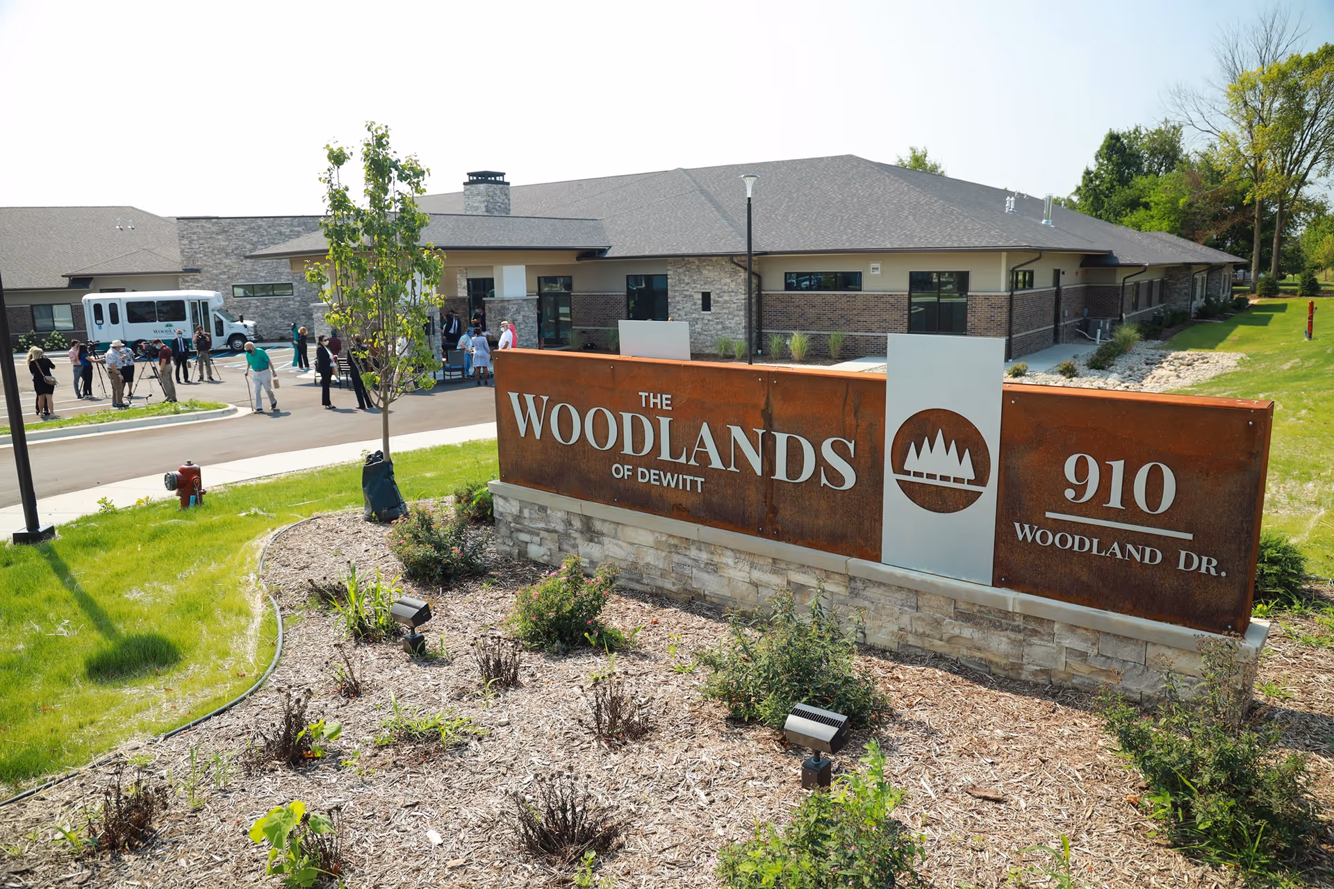Exterior view of The Woodlands of DeWitt senior living facility showing a large sign with the facility name and address 910 Woodland Dr. In the background, there is a modern building with people gathered near the entrance and a vehicle parked nearby. The area around the sign has landscaping with plants and mulch.