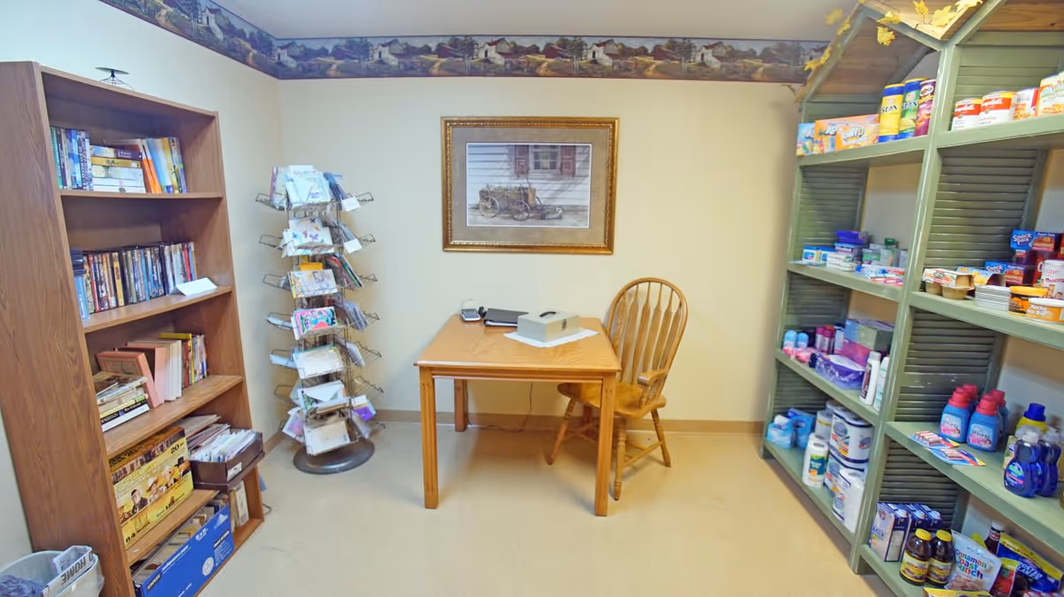 Small interior room with bookshelves and retail-style shelving stocked with household items, a table and chair, and a rotating card rack.