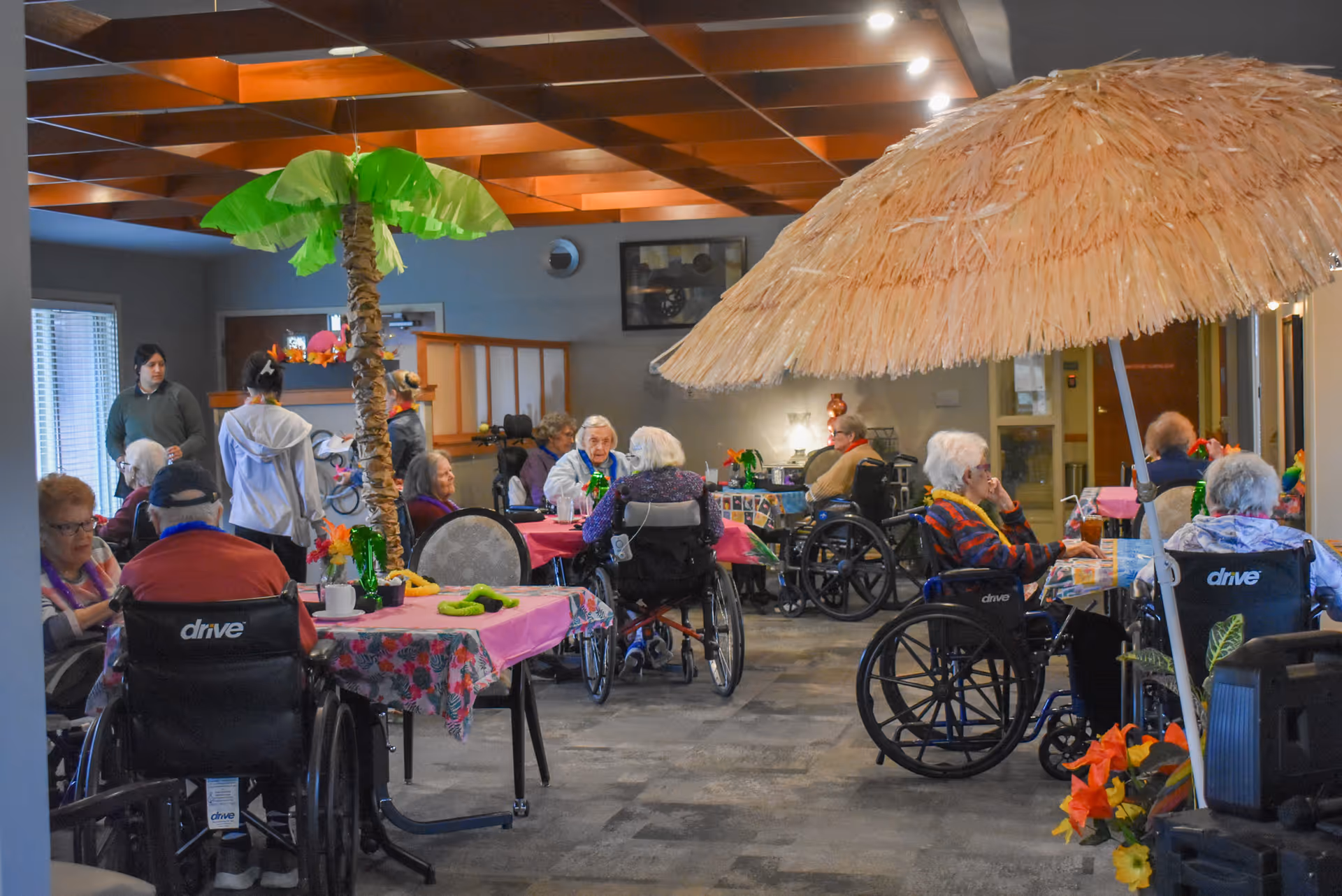 A group of elderly residents, many in wheelchairs, sitting around tables decorated with tropical-themed tablecloths and centerpieces under a thatched umbrella and a palm tree decoration in a common indoor area of a senior living facility.