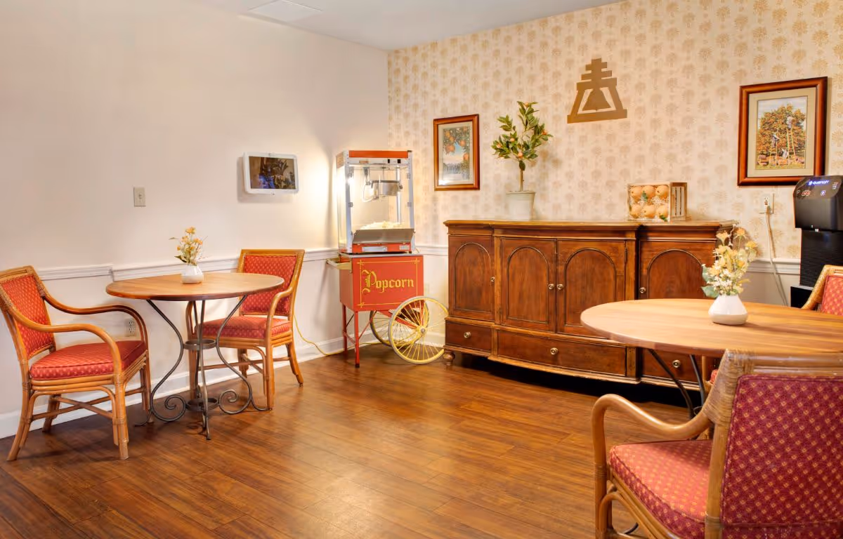 A cozy dining area with two round wooden tables, each surrounded by two wooden chairs with red cushioned seats. There is a vintage-style popcorn machine on wheels against the wall, next to a wooden sideboard with decorative items including a potted plant and framed artwork on the wallpapered wall behind it. The floor is wooden and the room has a warm, inviting atmosphere.