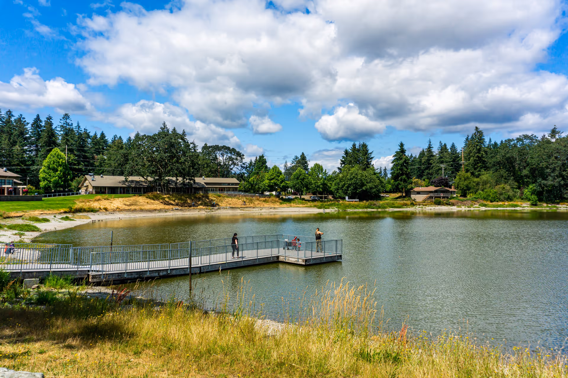 A scenic outdoor view of a lake with a metal pier extending into the water where three people are standing. Surrounding the lake are grassy areas, trees, and buildings under a partly cloudy blue sky.