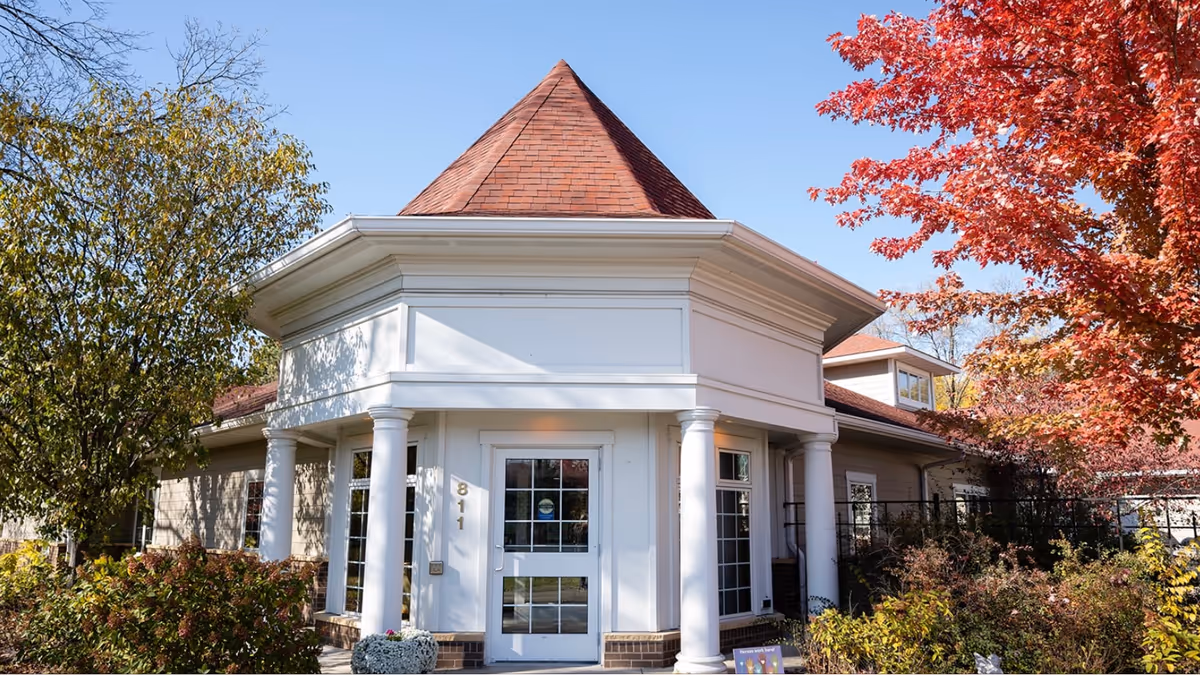 Front entrance of a white, columned senior living building with a red conical roof framed by autumn trees.
