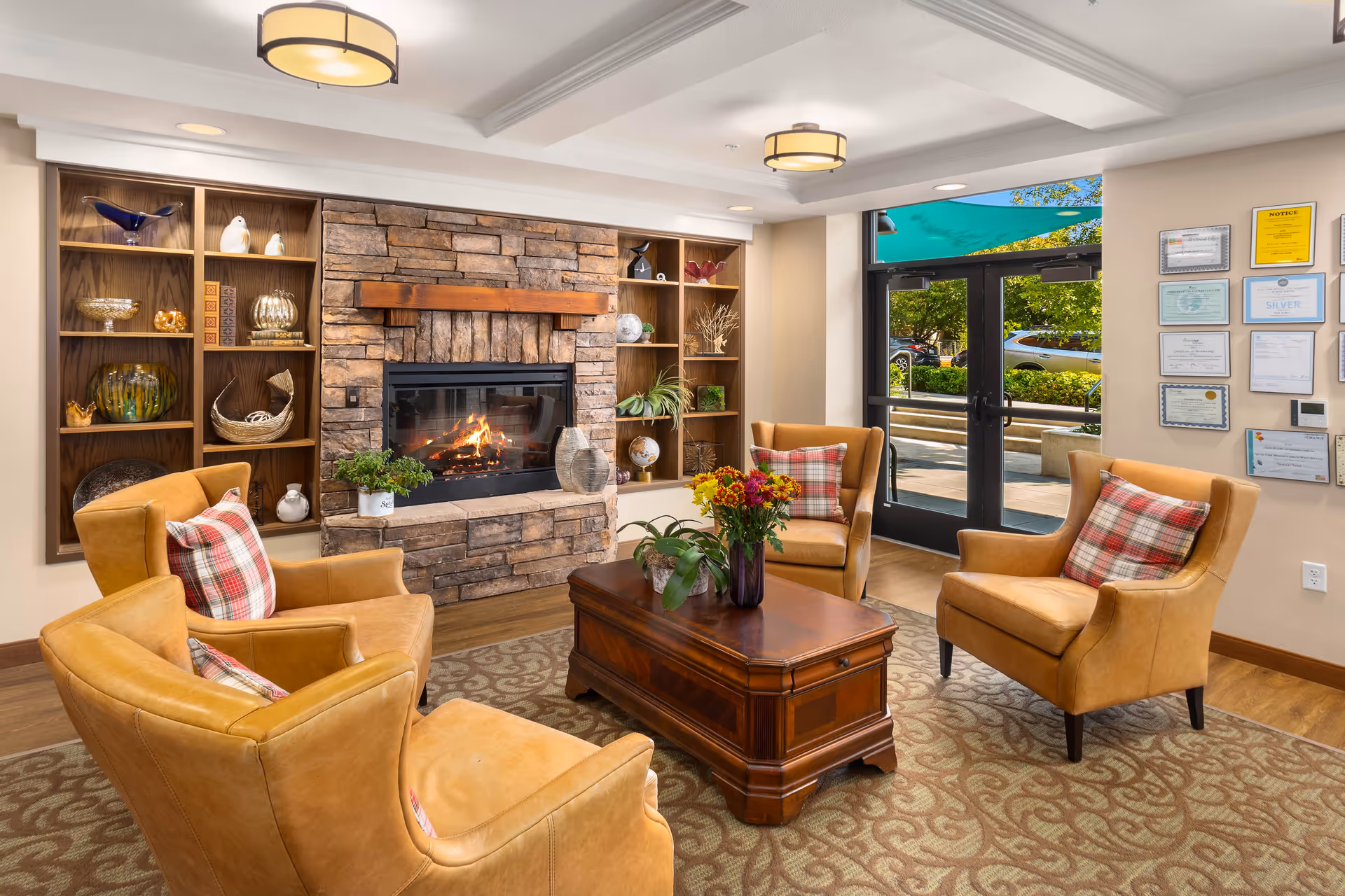 A cozy living room area with four tan leather armchairs arranged around a wooden coffee table with flowers and plants. Behind the chairs is a stone fireplace with a fire burning, flanked by built-in wooden shelves displaying decorative items. To the right, glass double doors open to an outdoor area with greenery and parked cars visible outside. The walls are adorned with framed certificates and notices.
