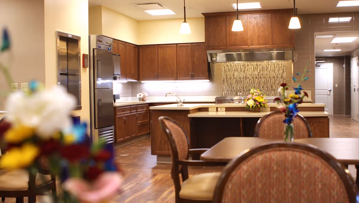 A well-lit kitchen area with wooden cabinets, a stainless steel refrigerator, and a stove with a decorative tile backsplash. In the foreground, there are tables and chairs with floral centerpieces, suggesting a dining or common area.