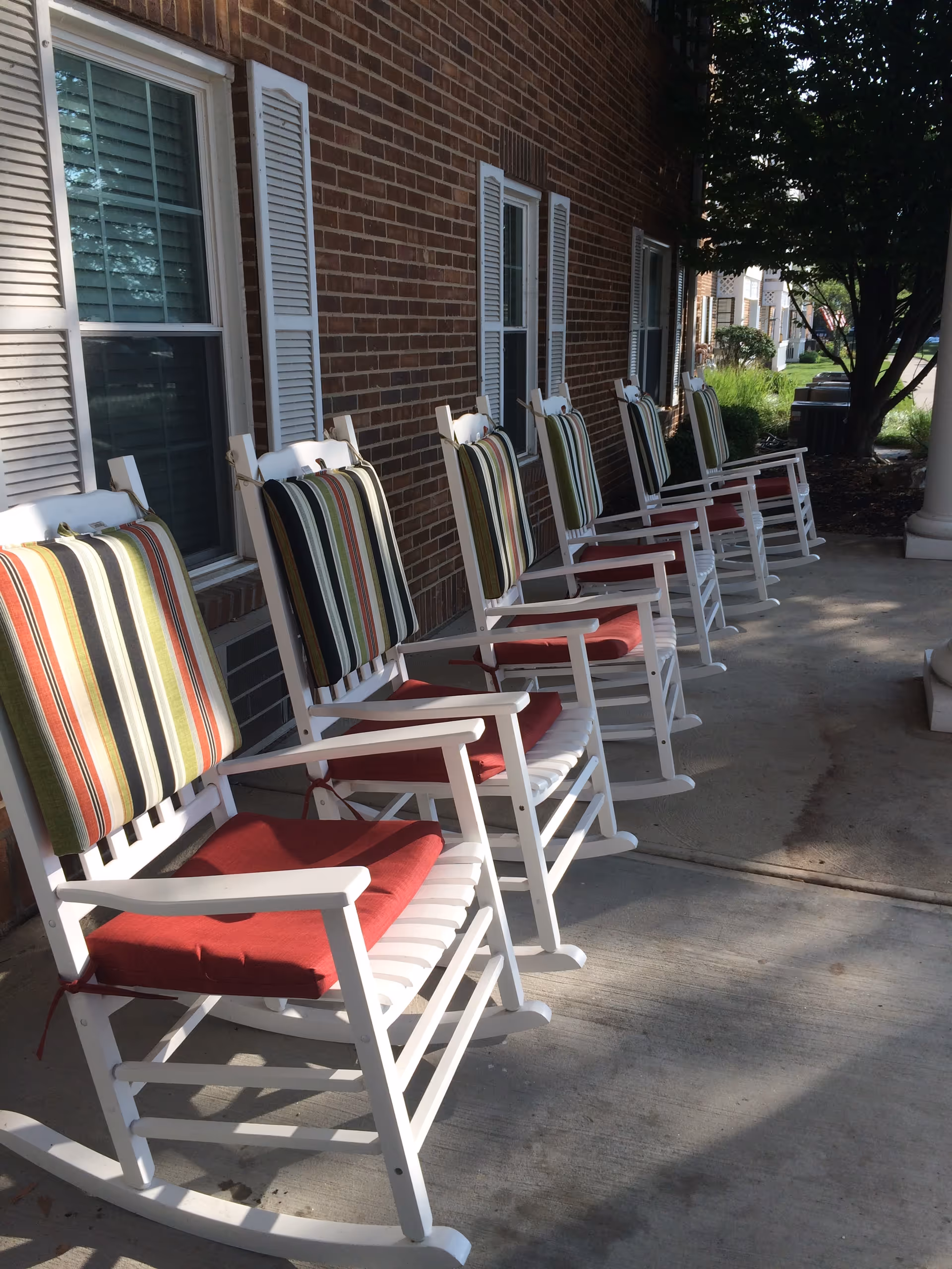 A row of white rocking chairs with striped back cushions and red seat pads lines a brick front porch.