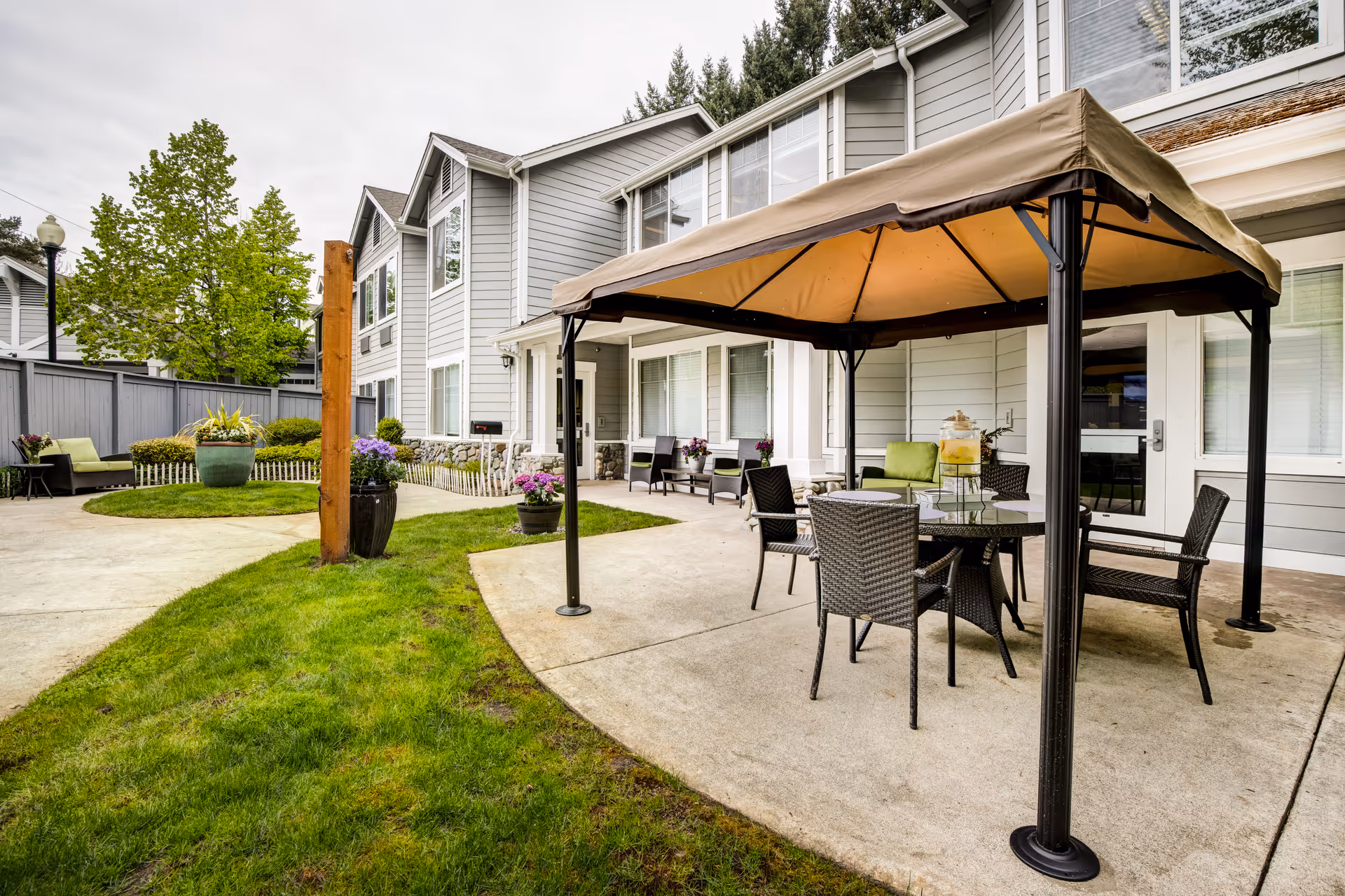 Outdoor patio area at Aegis Living Callahan House featuring a covered seating area with a table and four chairs, additional seating with green cushions, potted plants, and a well-maintained lawn surrounded by a gray fence and a two-story building with large windows.