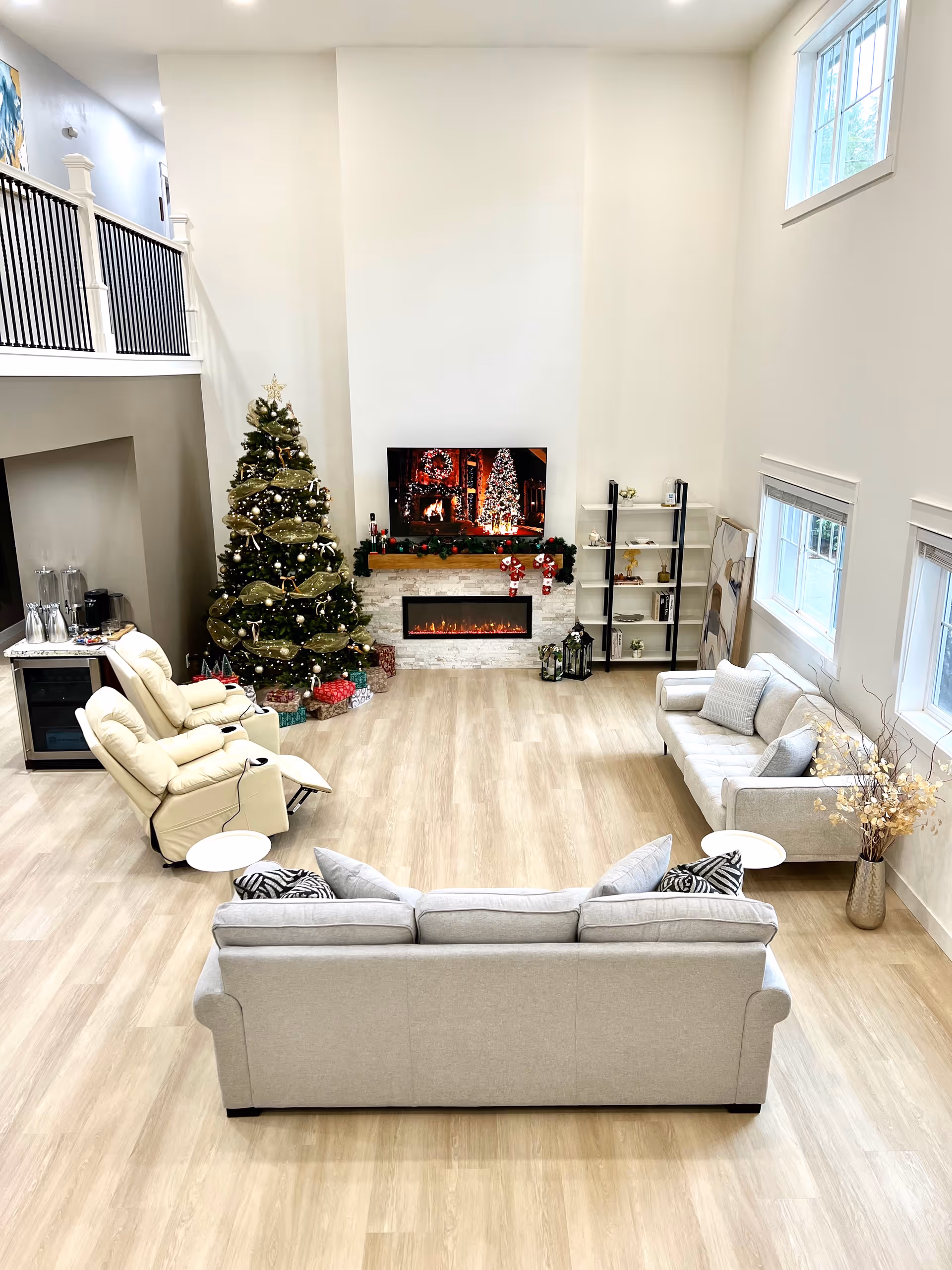 A spacious and bright living room decorated for Christmas with a large Christmas tree adorned with ornaments and lights, a modern electric fireplace with a TV above displaying a holiday scene, two beige reclining chairs, two light gray sofas with cushions, a tall shelving unit, and large windows letting in natural light.