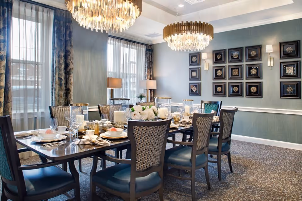 Elegant dining room with a long table set for a meal, featuring plates, glasses, napkins, and floral centerpieces. The room has large windows with sheer curtains and patterned drapes, two modern chandeliers hanging from the ceiling, and framed decorative art on the walls.