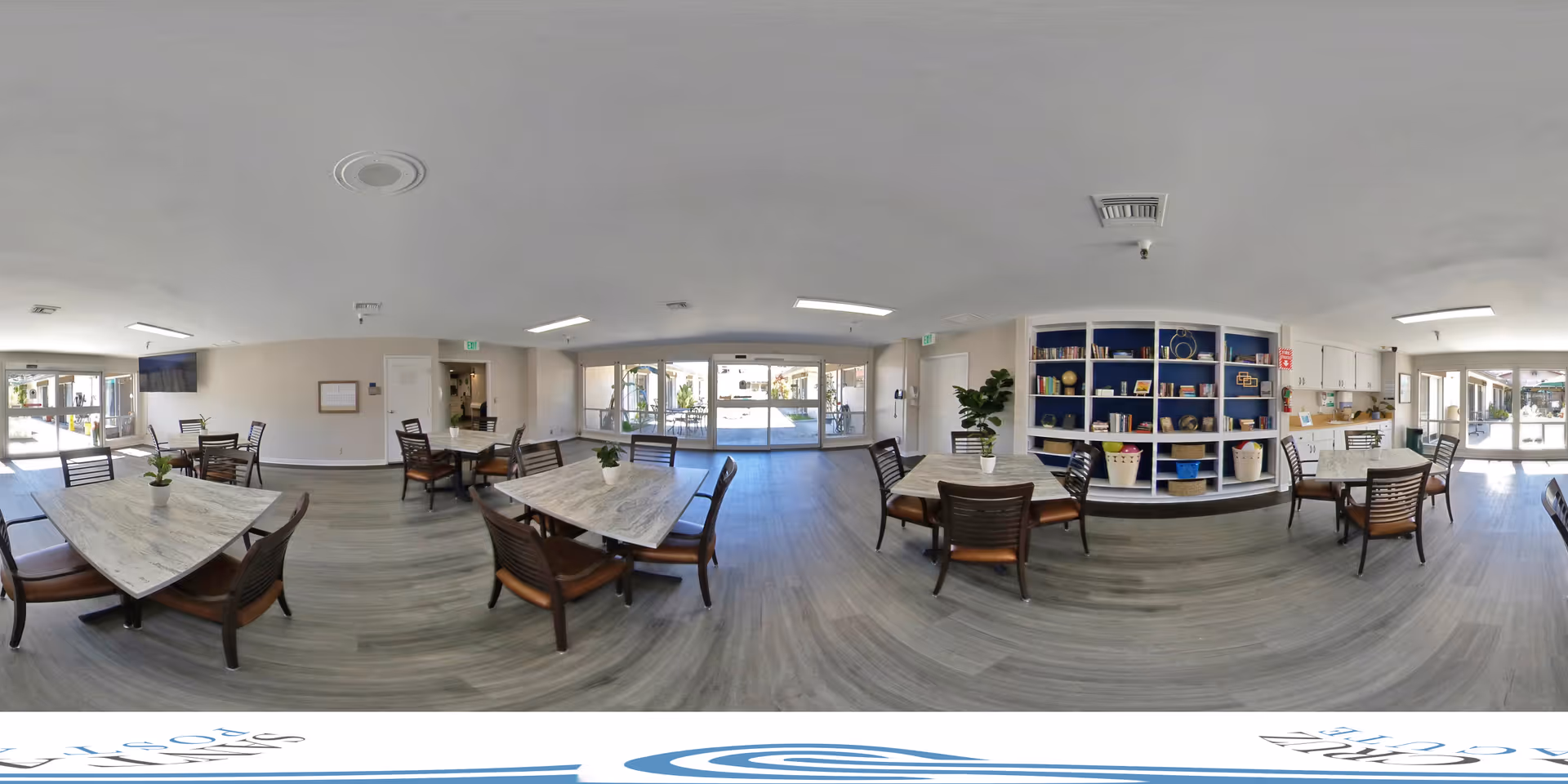 A spacious and well-lit common area with multiple tables and chairs arranged for seating. The room features large windows and glass doors allowing natural light to enter. There is a blue shelving unit with decorative items and baskets on the right side. The flooring is a light wood finish, and the walls are painted white. The area appears clean and inviting, suitable for social gatherings or activities.