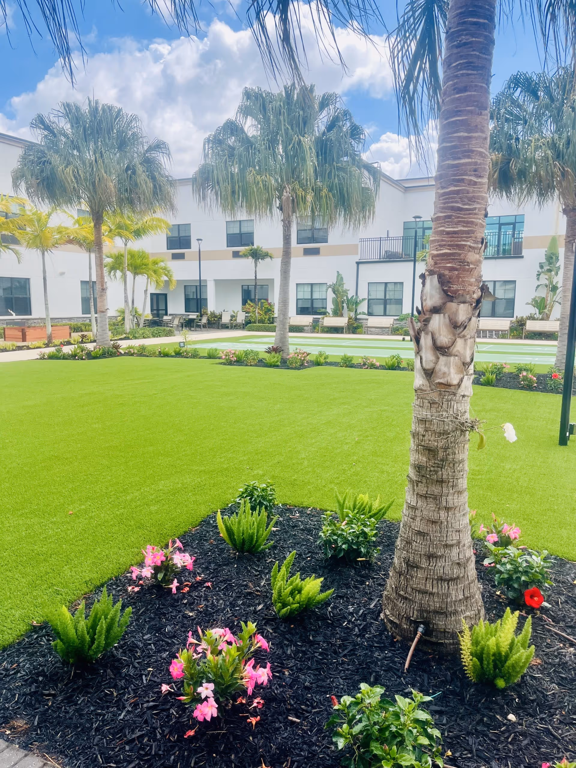 Well-manicured courtyard with palm trees, green lawn, flower beds, and a two-story building in the background.