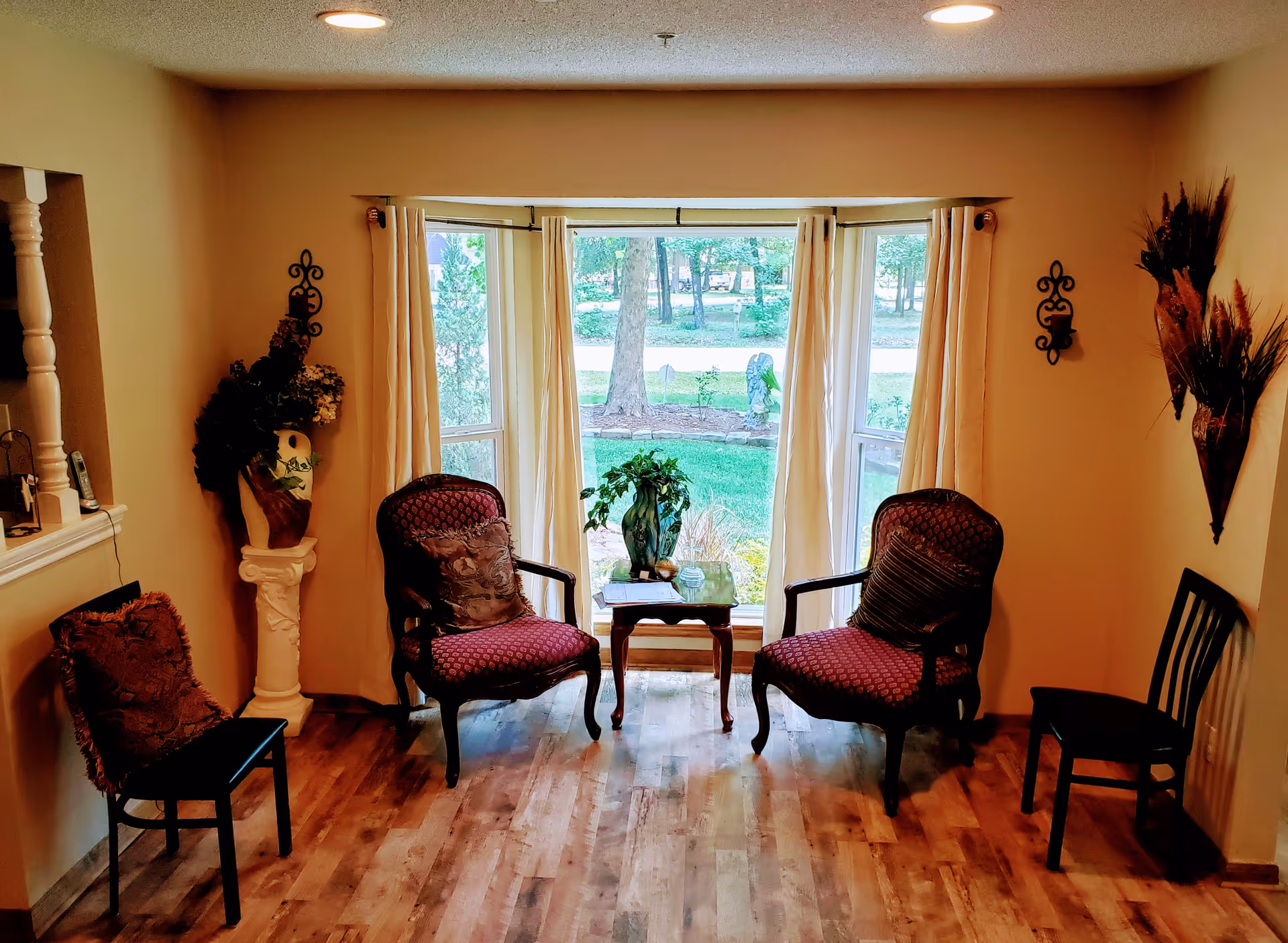 A cozy sitting area with four chairs arranged around a small wooden table holding a green plant. The room has wooden flooring, beige walls, and a large bay window with cream curtains that looks out onto a green lawn and trees. Decorative wall hangings and a floral arrangement on a pedestal add to the warm ambiance.