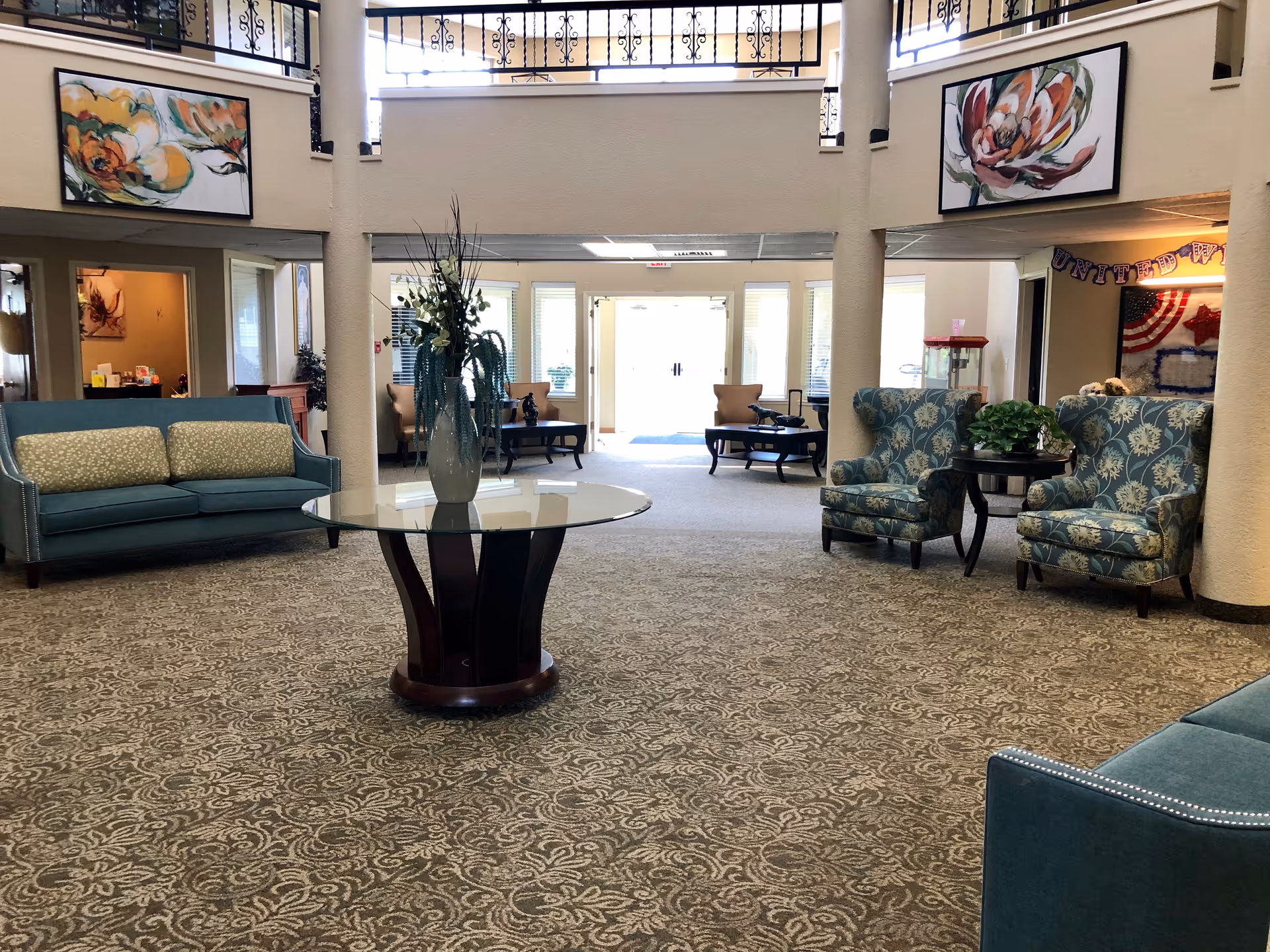Lobby of a senior living facility with sofas and armchairs around a central glass table topped with a vase, leading to double entry doors.