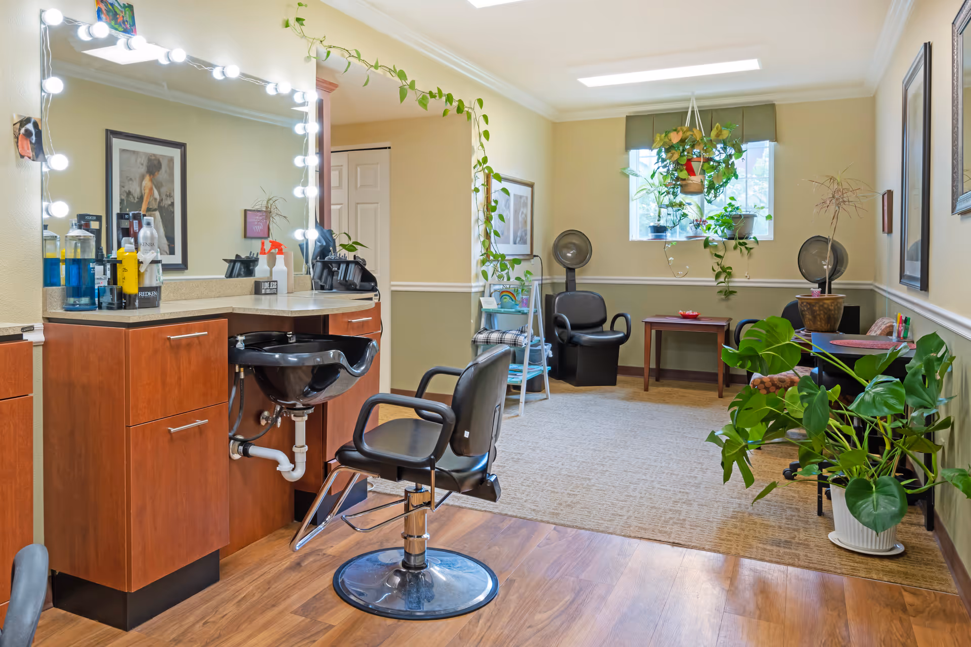 Interior of a hair salon area in a senior living facility with a black salon chair in front of a counter with a sink and a large mirror surrounded by lights. The room has wooden flooring near the salon area and carpeted flooring in the seating area with two black chairs under hair dryers, a small wooden table, and several potted plants including hanging plants near a window.